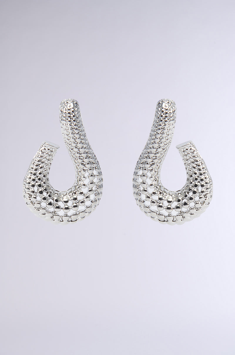 The DAY DREAMER SILVER EARRING showcases a looped design embellished with textured detailing and a reflective finish, set elegantly against a simple gray backdrop.