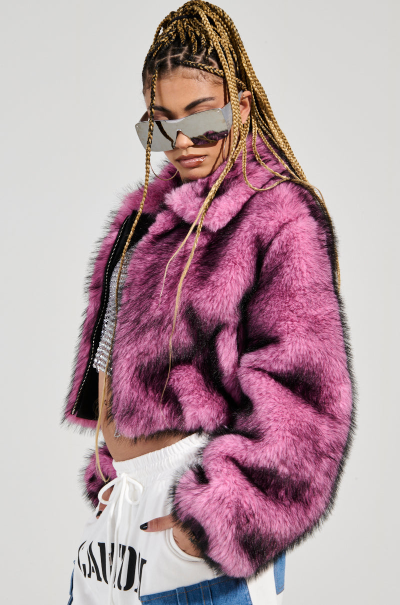 Dressed in the NOW YOU SEE ME FAUX FUR BOMBER IN PINK, an individual stands confidently with long braided hair and large, reflective sunglasses. They pair the vibrant jacket with white pants featuring black lettering, and a sparkly top glimmers from beneath the jacket, crafting a striking fashion look.