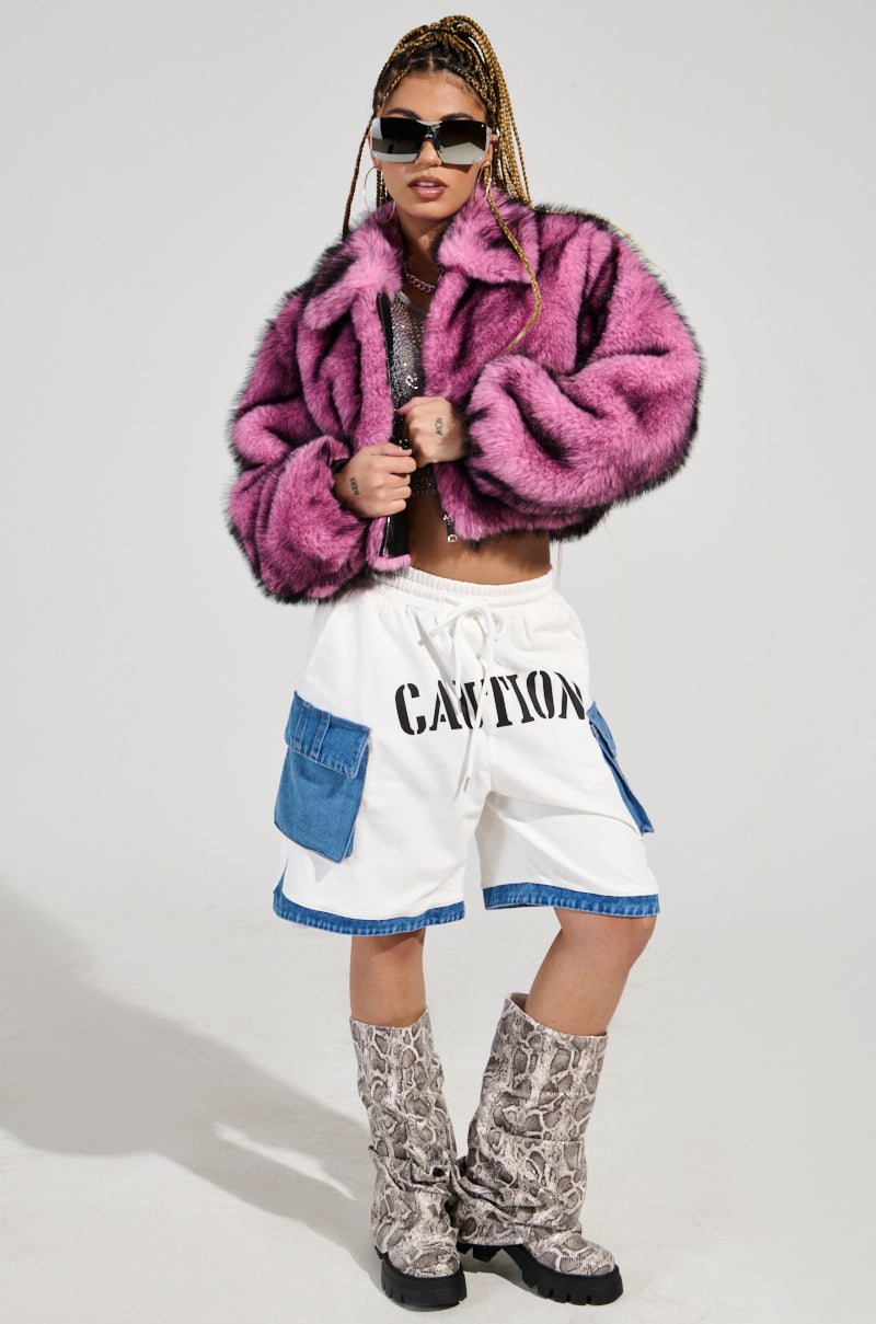 A person wearing the "Now You See Me" Faux Fur Bomber in a vibrant pink shade, paired with white shorts adorned with "CAUTION" lettering and blue denim accents, completes their look with tall snakeskin boots and sunglasses. They stand confidently against a plain background.