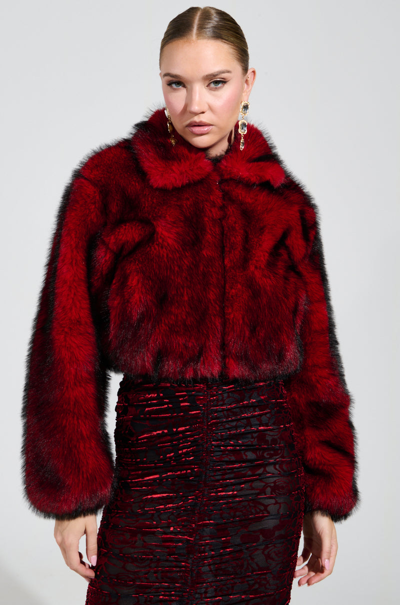 Dressed in the opulent NOW YOU SEE ME FAUX FUR BOMBER IN RED by AKIRA Label, paired with a dazzling red and black skirt, they present an elegant image. Their hair is impeccably styled, and they wear drop earrings while posing against a neutral backdrop.