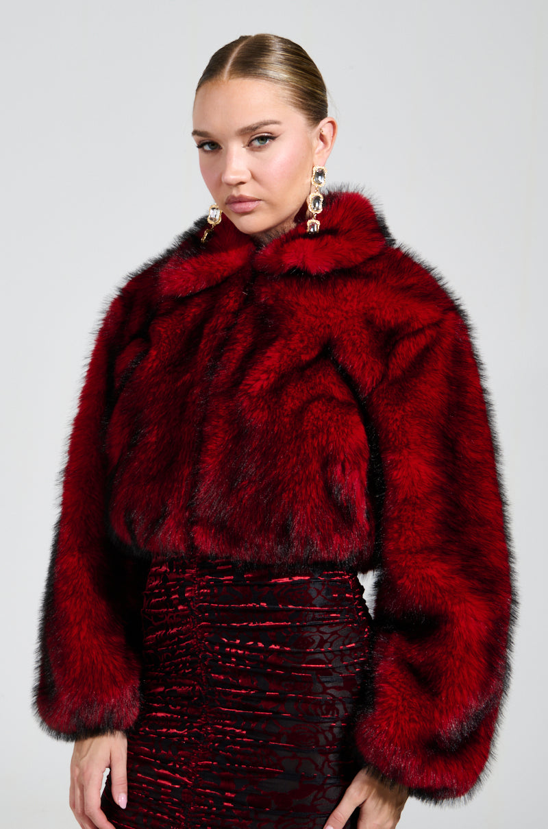 A person is dressed in the NOW YOU SEE ME FAUX FUR BOMBER IN RED by AKIRA Label, paired with a matching textured skirt. Their sleek, pulled-back hair is complemented by large, decorative earrings against a plain, light-colored background.