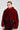 A person is dressed in the NOW YOU SEE ME FAUX FUR BOMBER IN RED by AKIRA Label, paired with a matching textured skirt. Their sleek, pulled-back hair is complemented by large, decorative earrings against a plain, light-colored background.