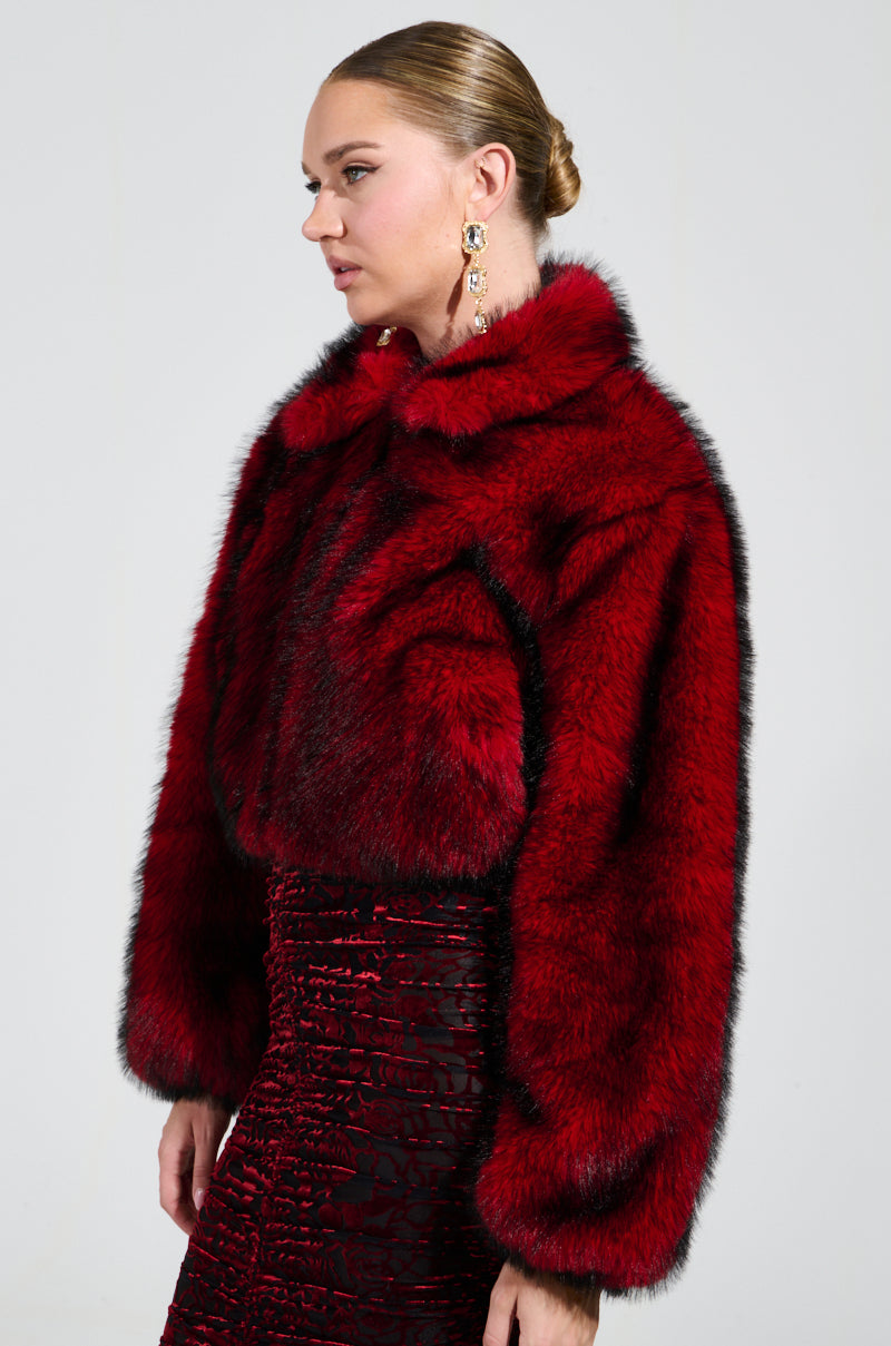 A woman with her hair tied back is dressed in a luxurious NOW YOU SEE ME FAUX FUR BOMBER IN RED from the AKIRA Label, featuring black accents and a matching skirt. She accessorizes with large, elaborate earrings and stands against a plain gray background.