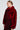 A woman with her hair tied back is dressed in a luxurious NOW YOU SEE ME FAUX FUR BOMBER IN RED from the AKIRA Label, featuring black accents and a matching skirt. She accessorizes with large, elaborate earrings and stands against a plain gray background.