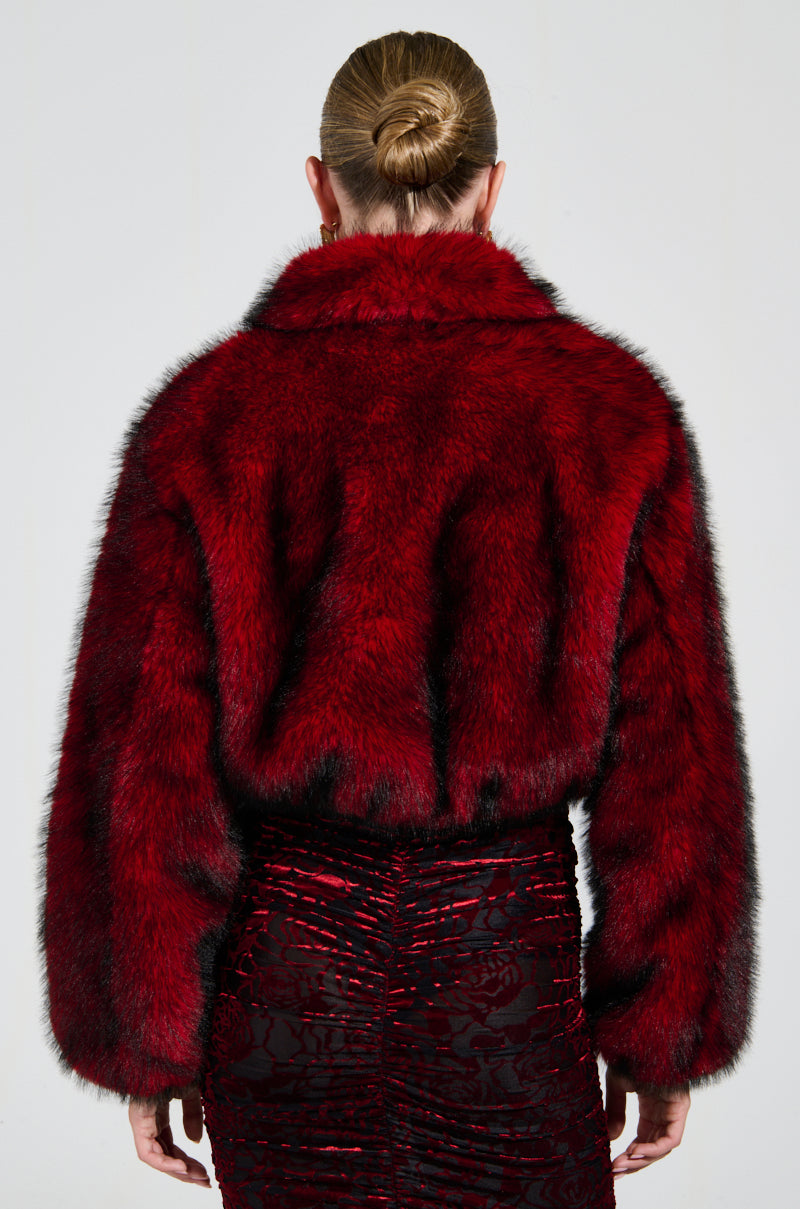 A person with a bun is wearing the NOW YOU SEE ME FAUX FUR BOMBER IN RED from AKIRA Label and a black skirt featuring red detailing, depicted from the back against a plain background.