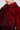 A person draped in the NOW YOU SEE ME FAUX FUR BOMBER IN RED, featuring a luxurious faux fur collar, styled with large ornate gold earrings embellished with jewels. The bomber jacket exhibits a textured, shiny appearance against a neutral gray backdrop.