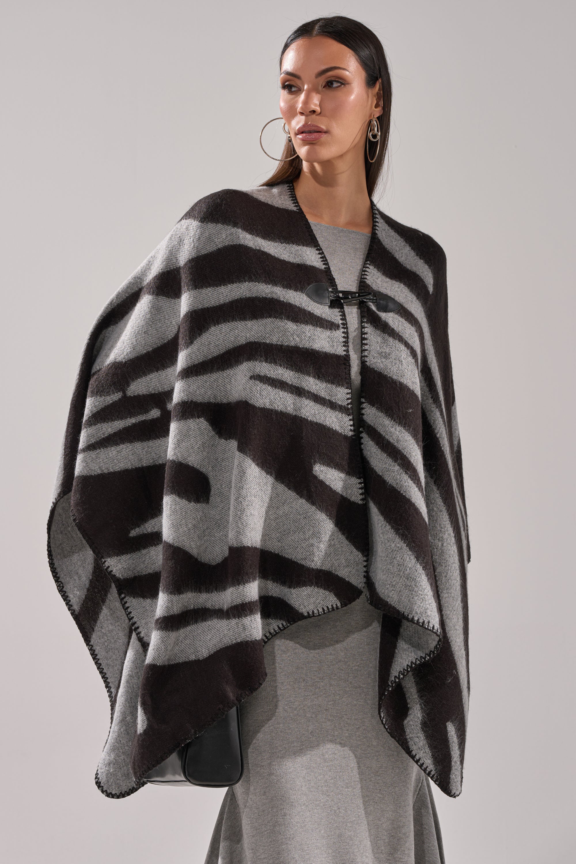 A woman models the WILD CHILD KNIT PONCHO, featuring an oversized black and gray zebra print, layered over a light gray outfit. She stands against a plain background, gazing to the side with a neutral expression. 1
