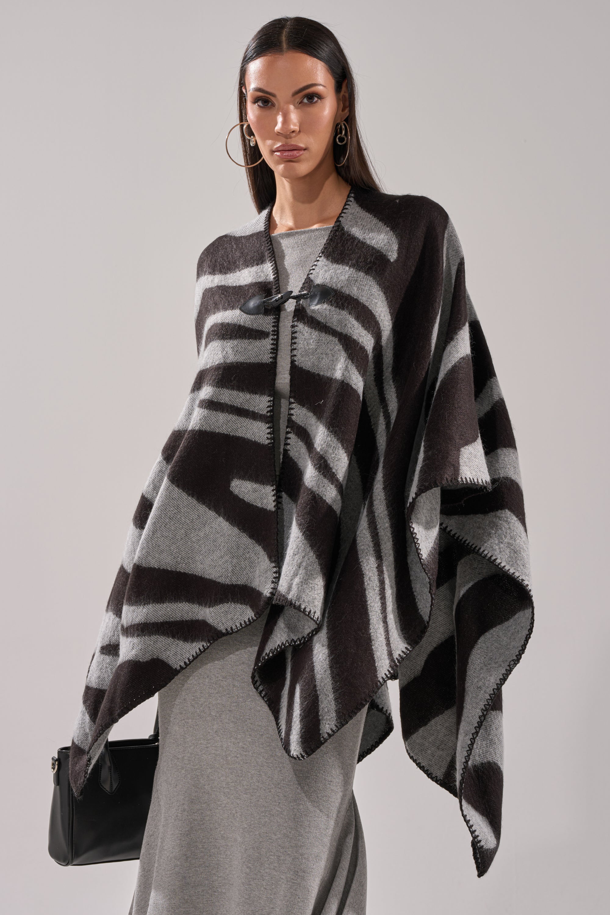 A woman wearing the WILD CHILD KNIT PONCHO in black and gray over a gray dress, with large hoop earrings and a black handbag, stands against a plain light background. 0