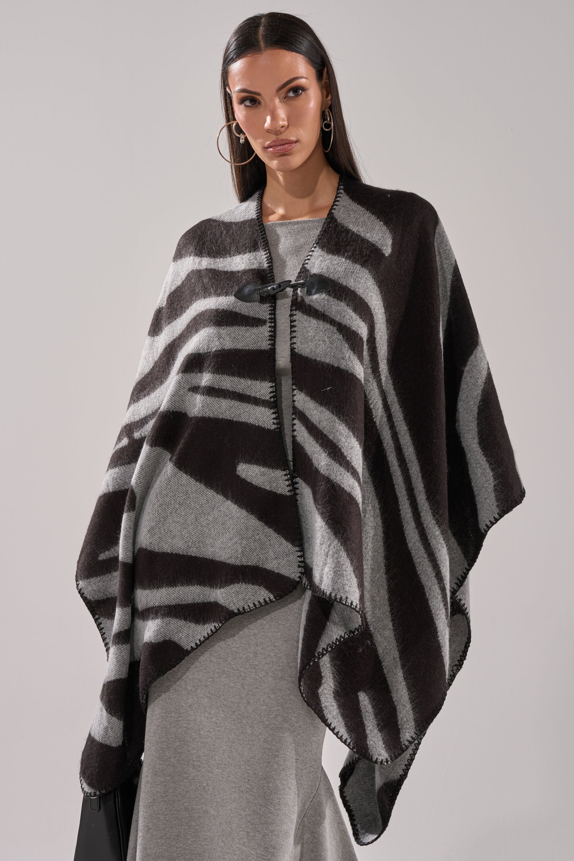 A woman with long dark hair wears the WILD CHILD KNIT PONCHO in black and gray zebra print over a light gray dress, paired with hoop earrings. She stands against a plain light background with a neutral expression. 2