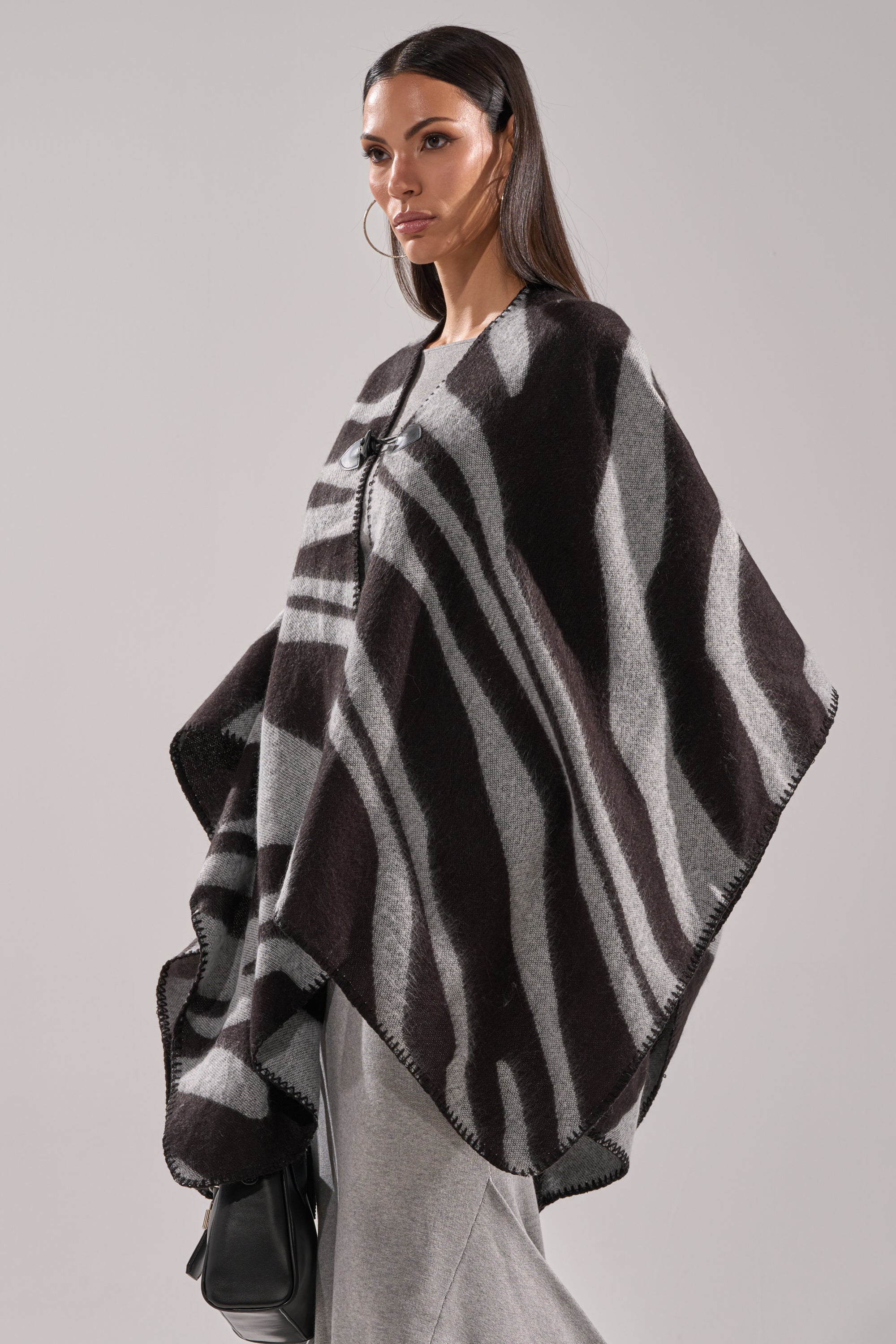 A woman with long dark hair wears the WILD CHILD KNIT PONCHO, showcasing its oversized black and gray zebra print over gray pants. She carries a black bag and poses against a plain light background. 3