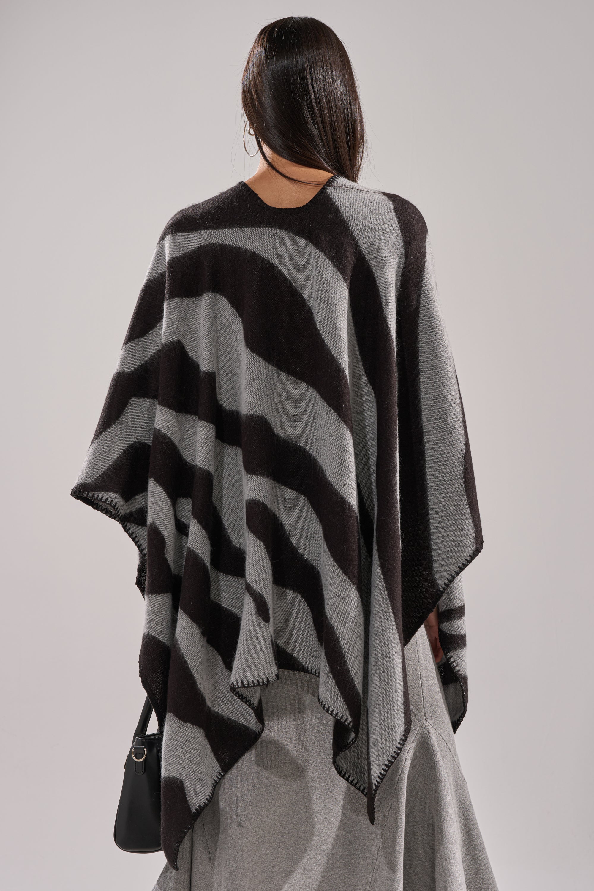 A person with long, straight brown hair is seen from behind, wearing the WILD CHILD KNIT PONCHO—black and gray with a zigzag hem and oversized silhouette—paired with a light gray skirt and holding a black handbag. 4