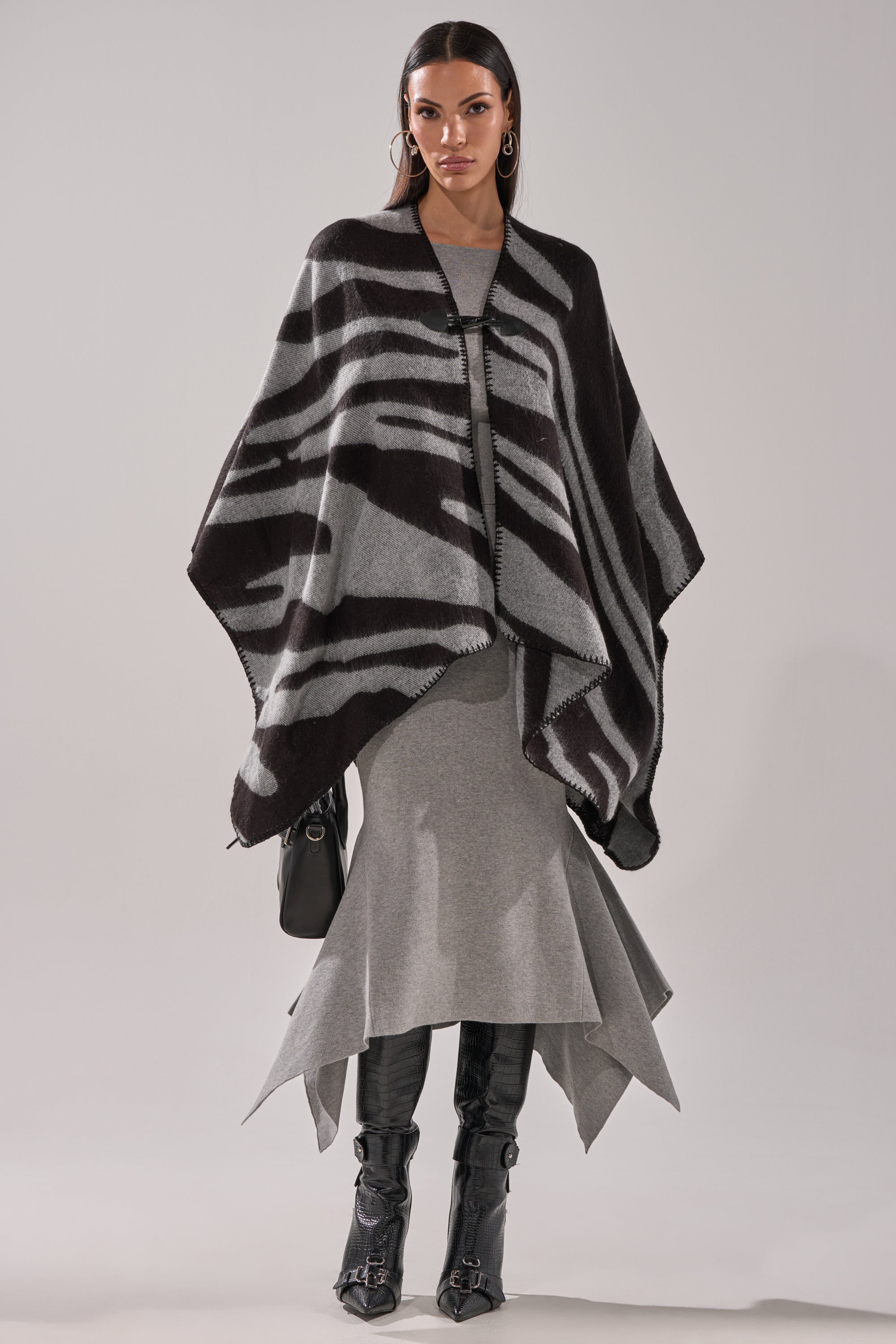 A woman faces forward, wearing the WILD CHILD KNIT PONCHO with a zebra print in an oversized fit, paired with a gray asymmetrical-hem dress, knee-high black boots, large hoop earrings, and carrying a black handbag. Her expression is neutral. 5