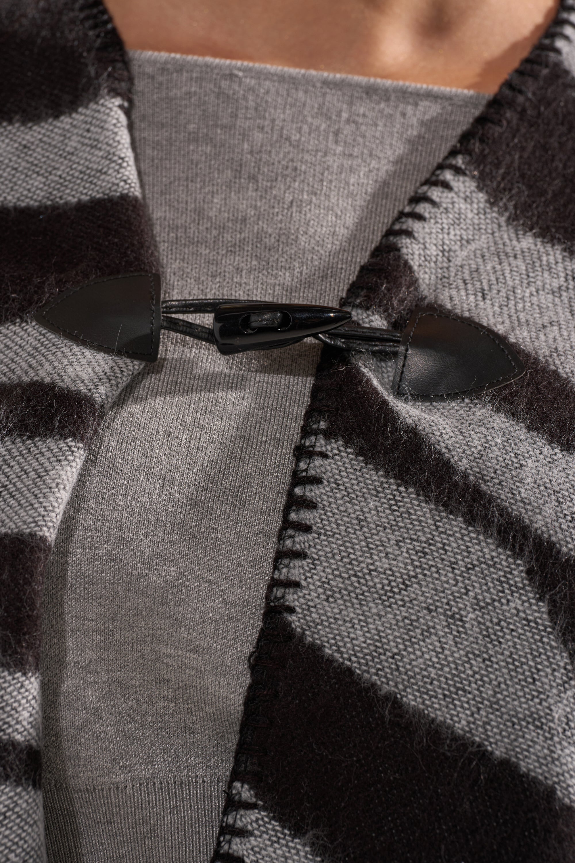 Close-up of the WILD CHILD KNIT PONCHO, featuring bold black and gray stripes, an oversized silhouette, and a black leather toggle clasp over a light gray knit top, showcasing its textured knit and detailed stitching. 6