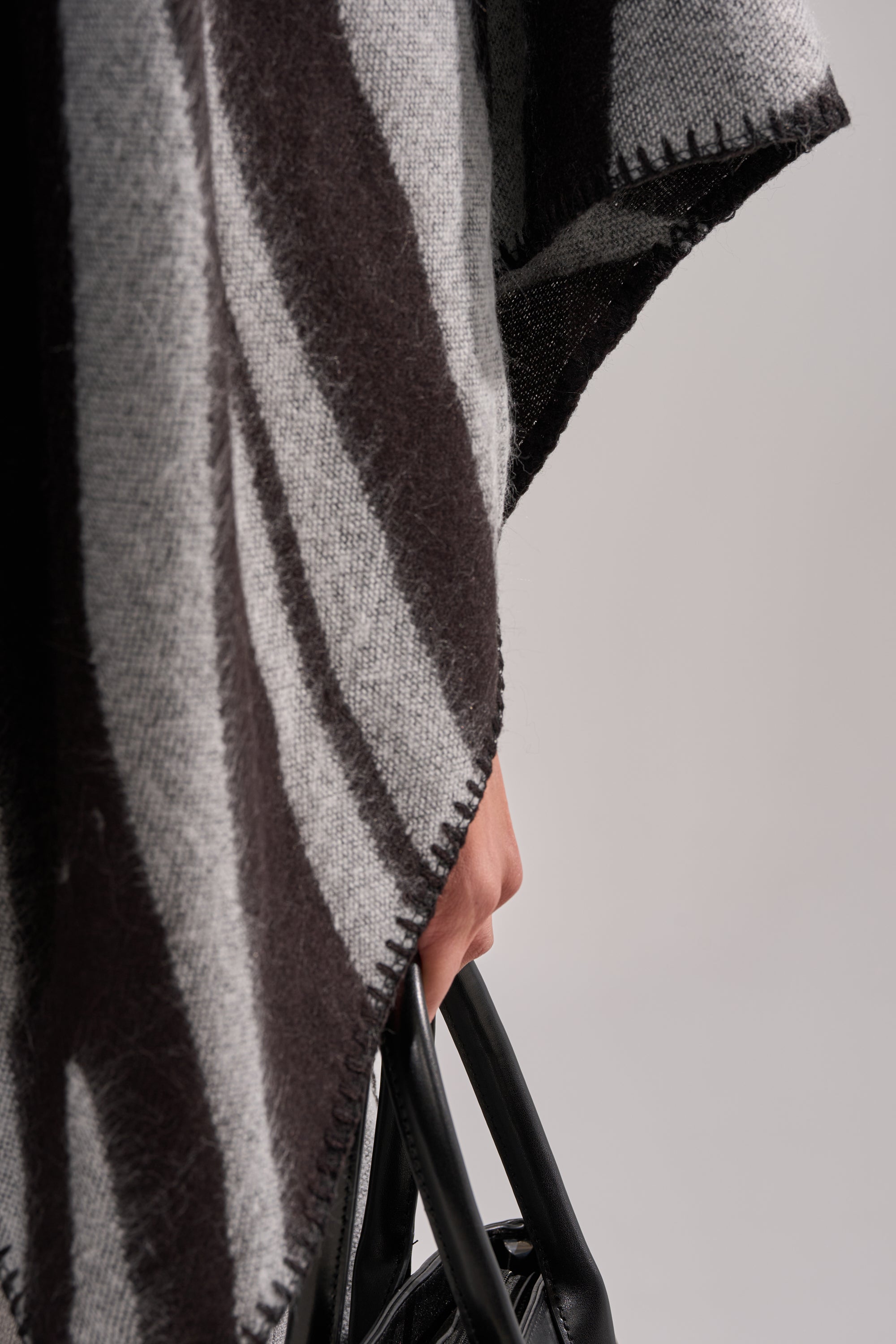 Close-up of a person’s hand holding a black handbag while the WILD CHILD KNIT PONCHO, in gray and black, is draped over their shoulder against a plain light background. 7