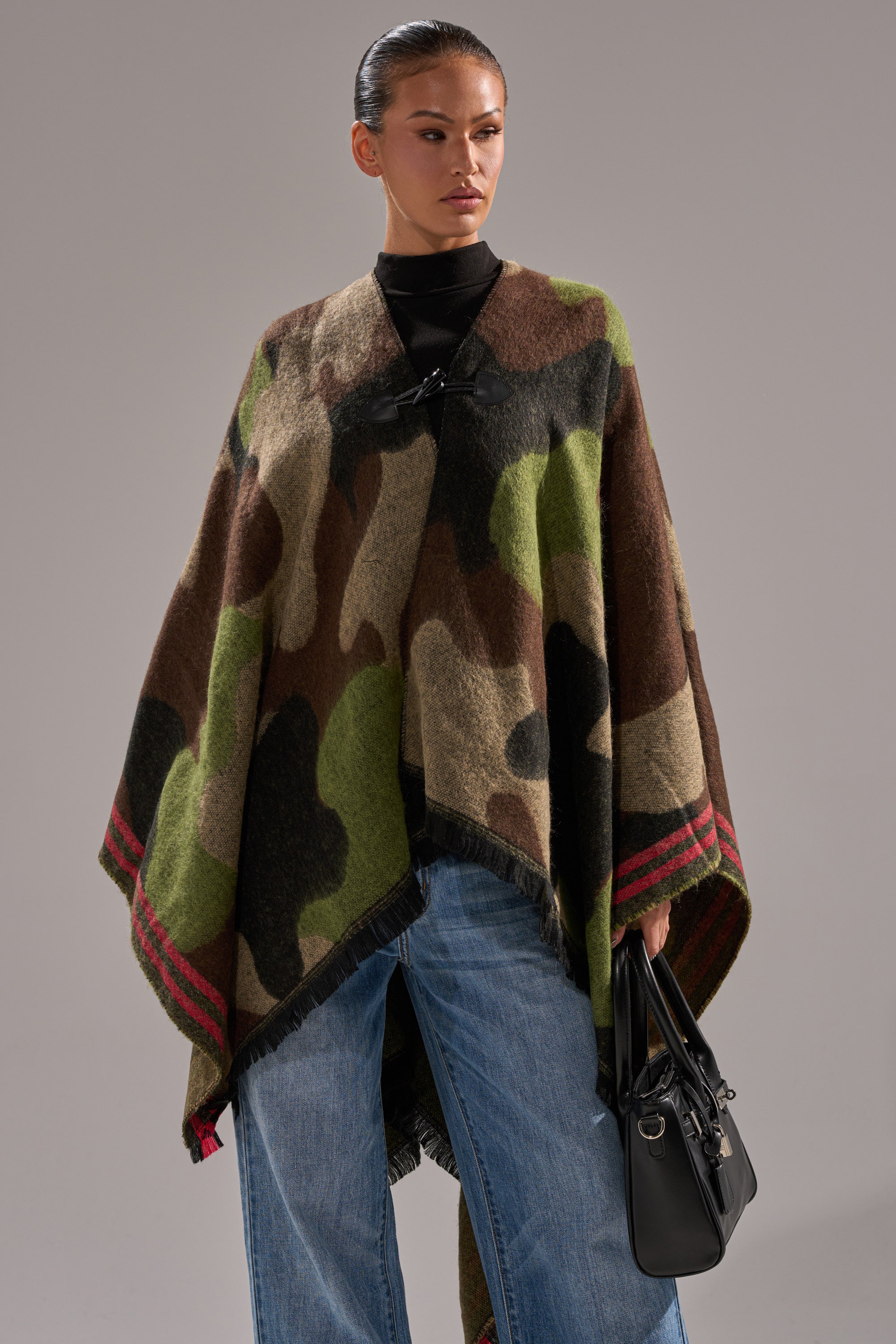 Model wears the YOU CAN'T SEE ME CAMO PONCHO with red-edged stripes, wide-leg blue jeans, and a black handbag, posing against a plain gray background. 1