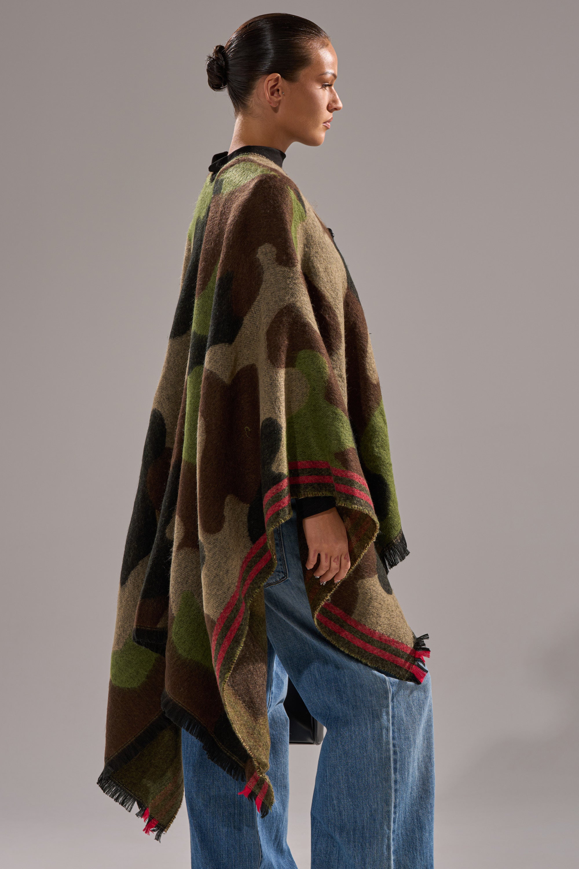 A person stands in profile wearing the YOU CAN'T SEE ME CAMO PONCHO with red accents over blue jeans. Their hair is tied back in a neat bun, set against a plain, light grey background. 2