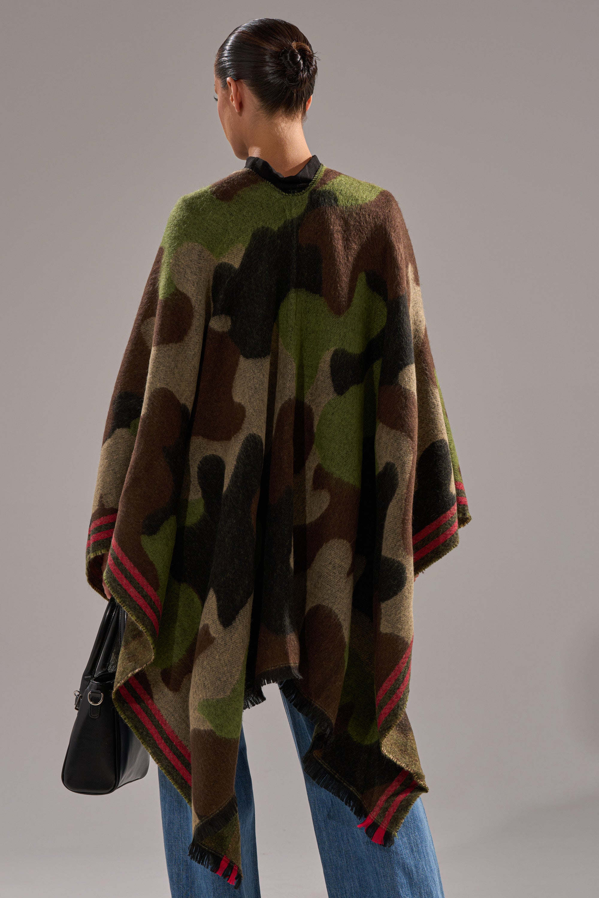 A person with dark hair in a bun models the YOU CAN'T SEE ME CAMO PONCHO with red-striped edges, styled with blue jeans and a black bag, standing against a plain gray background—ideal for bonfire nights. 3