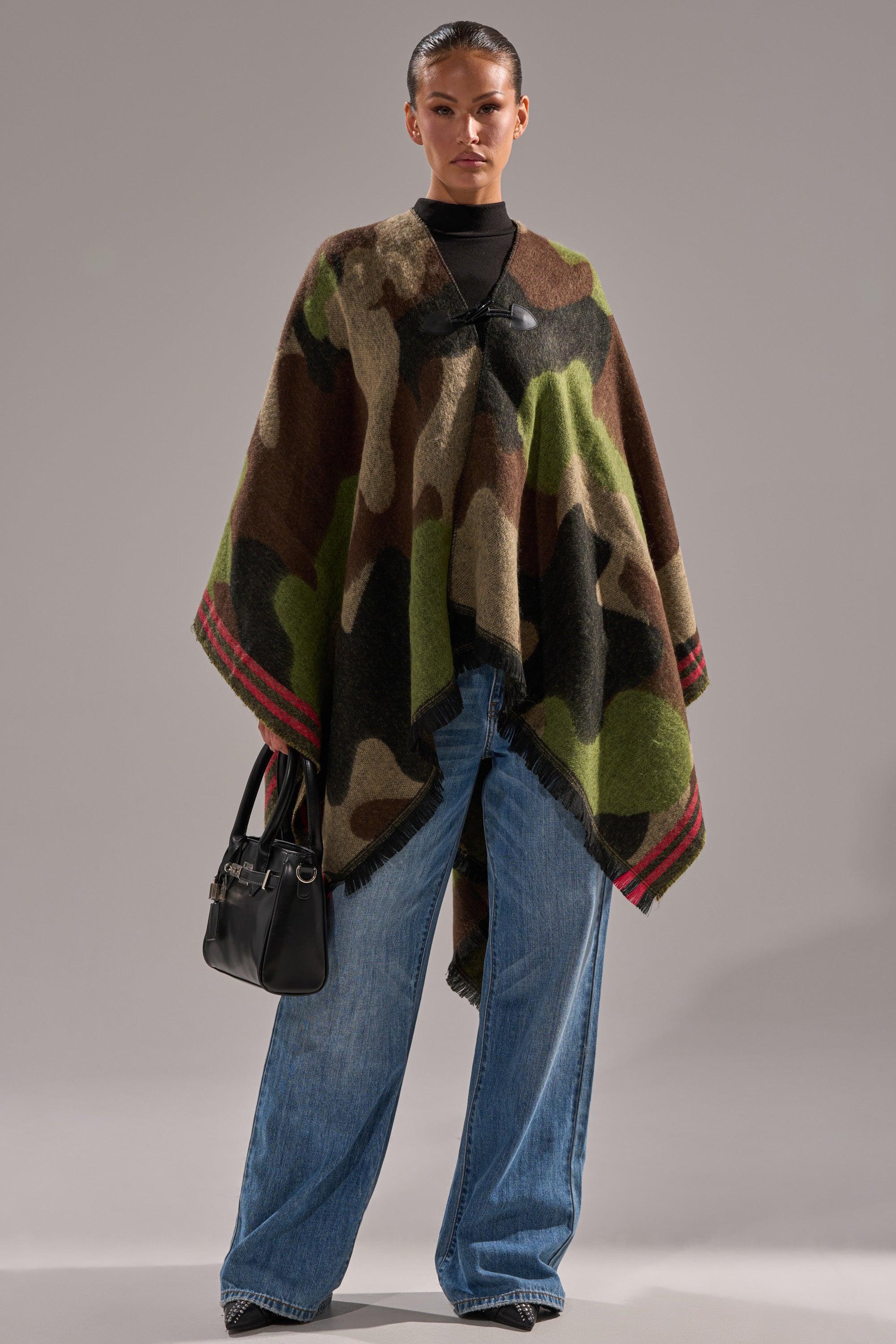 A woman models the YOU CAN'T SEE ME CAMO PONCHO, featuring soft knit fabric and red trim, styled with a black turtleneck and blue wide-leg jeans—an ideal bonfire accessory for cool evenings. 4