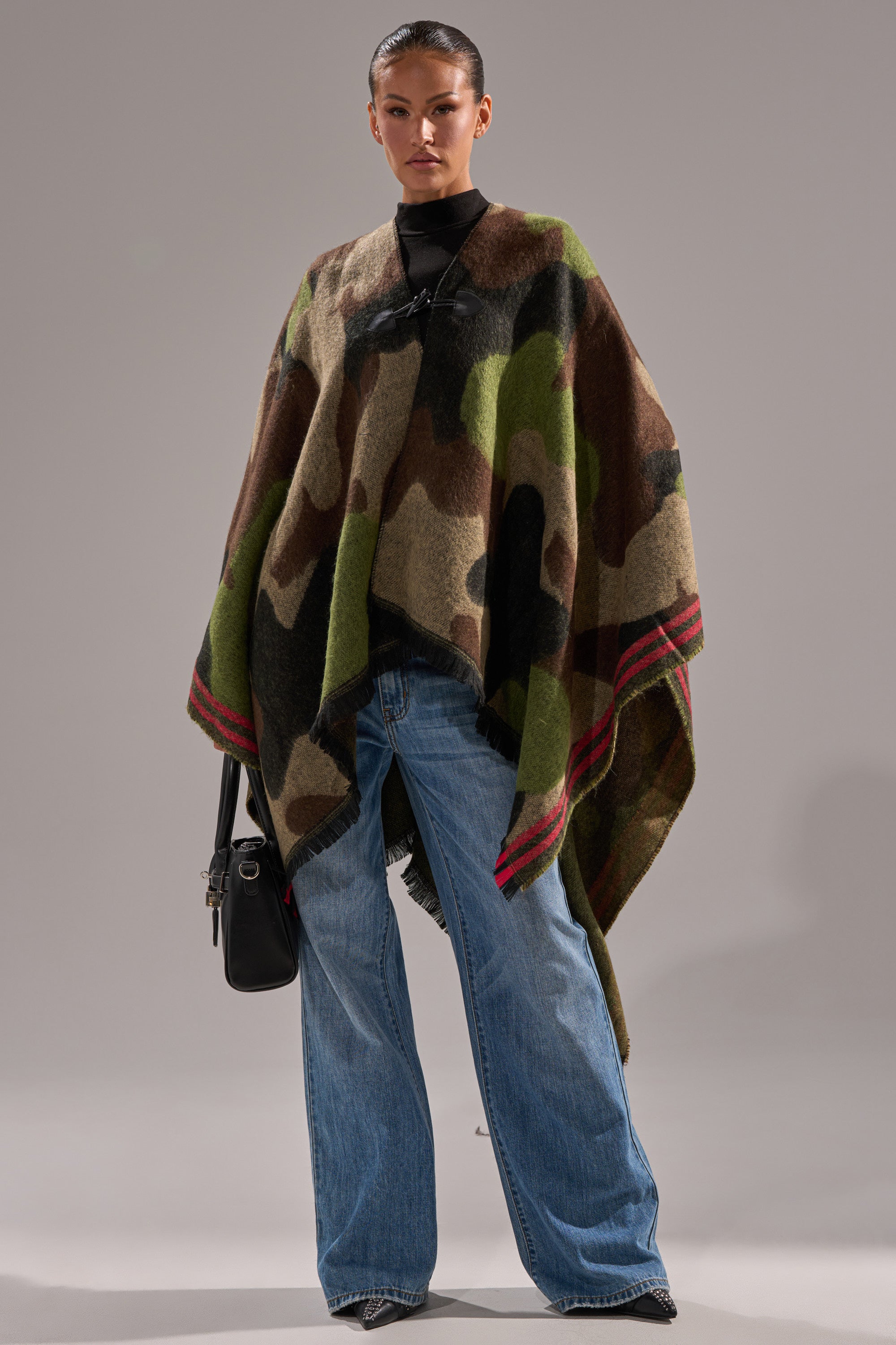 A woman stands facing forward, wearing the YOU CAN'T SEE ME CAMO PONCHO with red-edged stripes, blue wide-leg jeans, black pointed shoes, and a black handbag—her hair slicked back for a cozy bonfire accessory vibe. 5