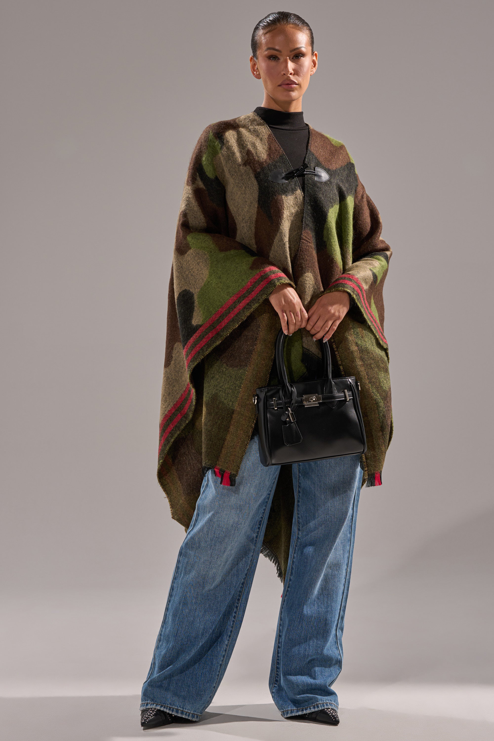 A woman wears the YOU CAN'T SEE ME CAMO PONCHO with red trim over a black top and wide-leg blue jeans, holding a black handbag. This soft knit poncho is the perfect bonfire accessory, set against a plain gray background. 6