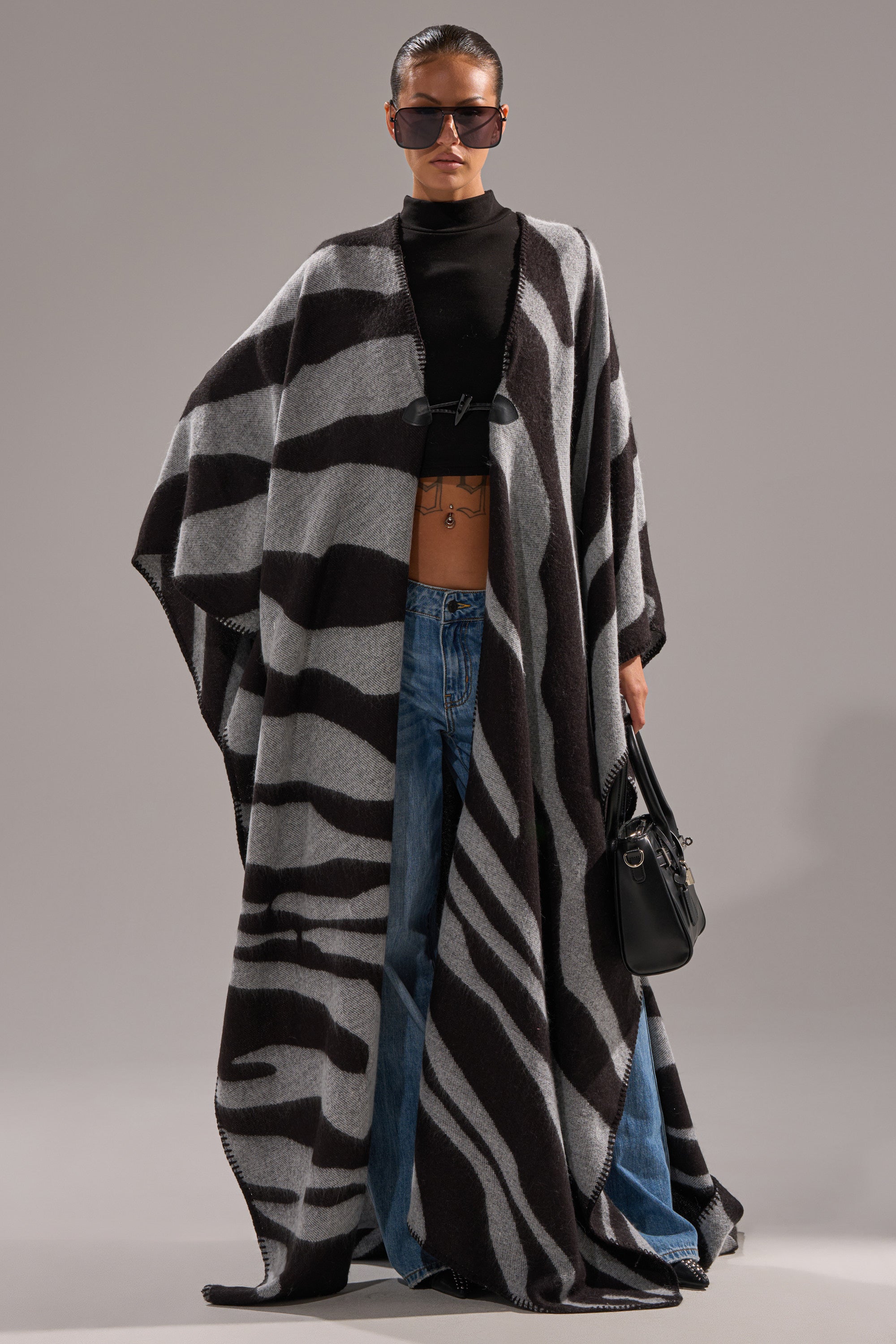 A person wears oversized blue jeans, a black crop top, large sunglasses, a black handbag, and the WILD ONE KNIT MAXI PONCHO, standing confidently against a gray background.