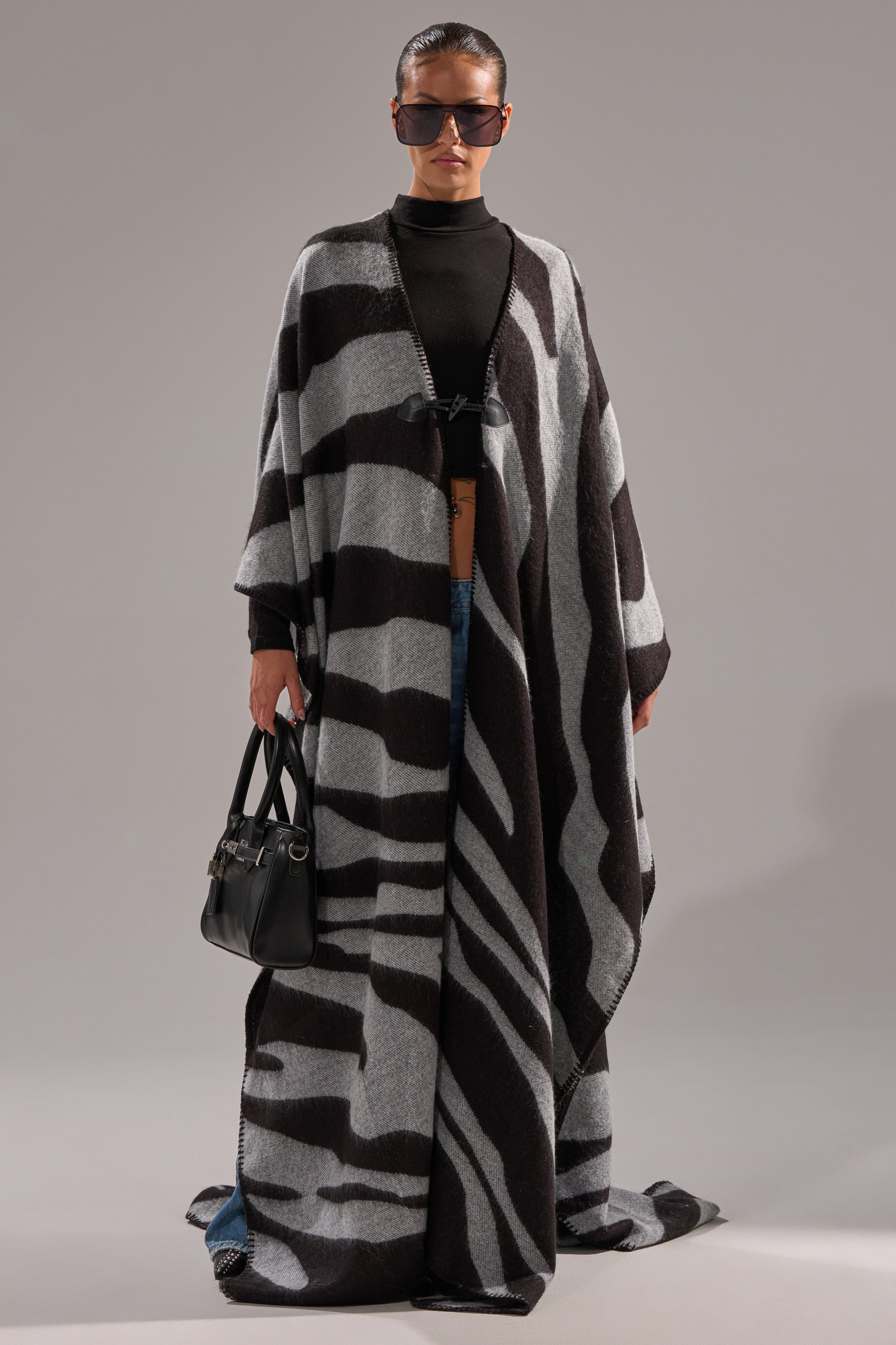 A person stands against a plain background wearing sunglasses, jeans, a black top, and the WILD ONE KNIT MAXI PONCHO—a long, flowing zebra-striped cape—while holding a black handbag.