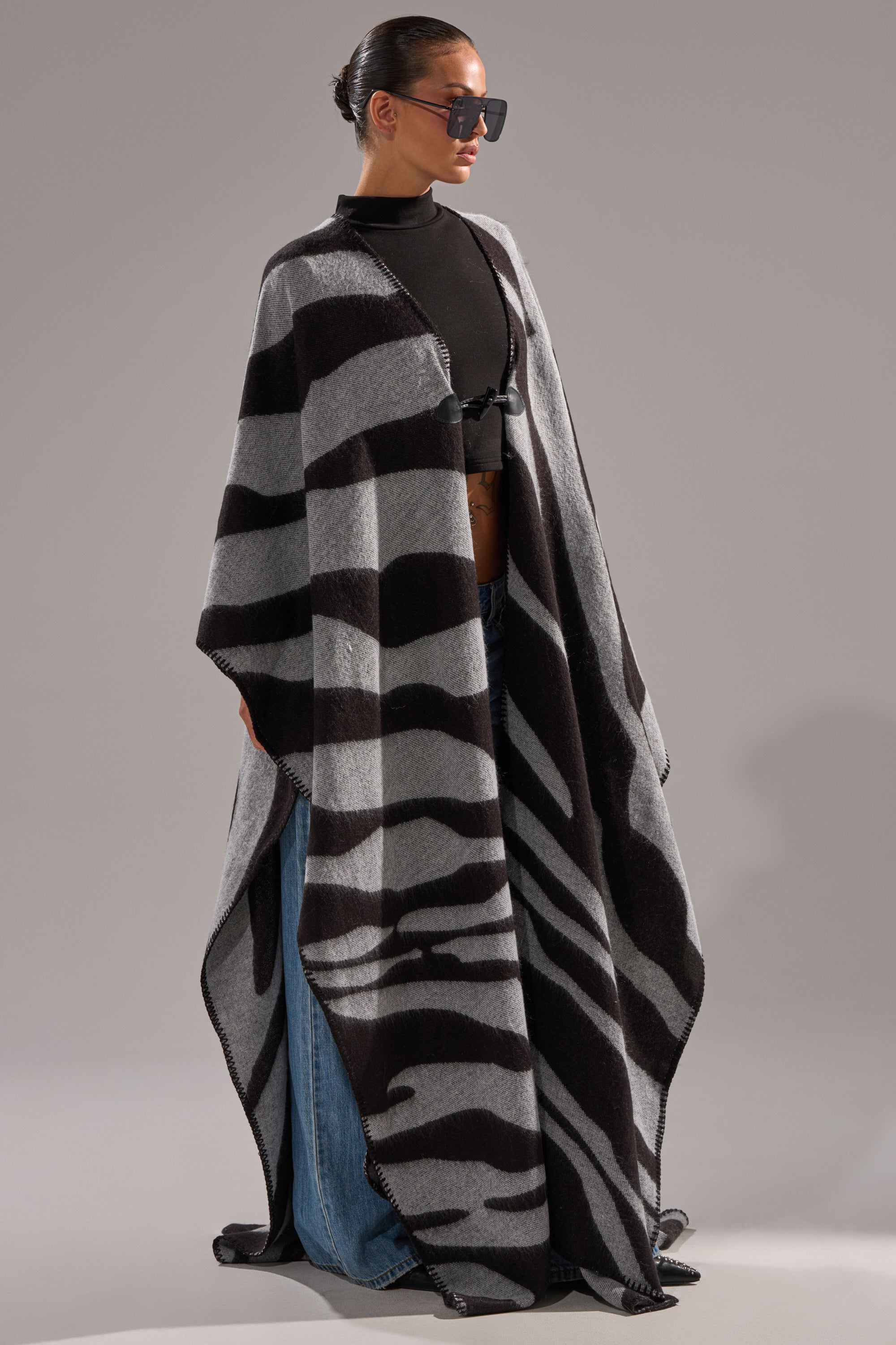 A person wearing sunglasses, a black top, blue jeans, and the WILD ONE KNIT MAXI PONCHO with black-and-gray zebra stripes stands against a plain background.