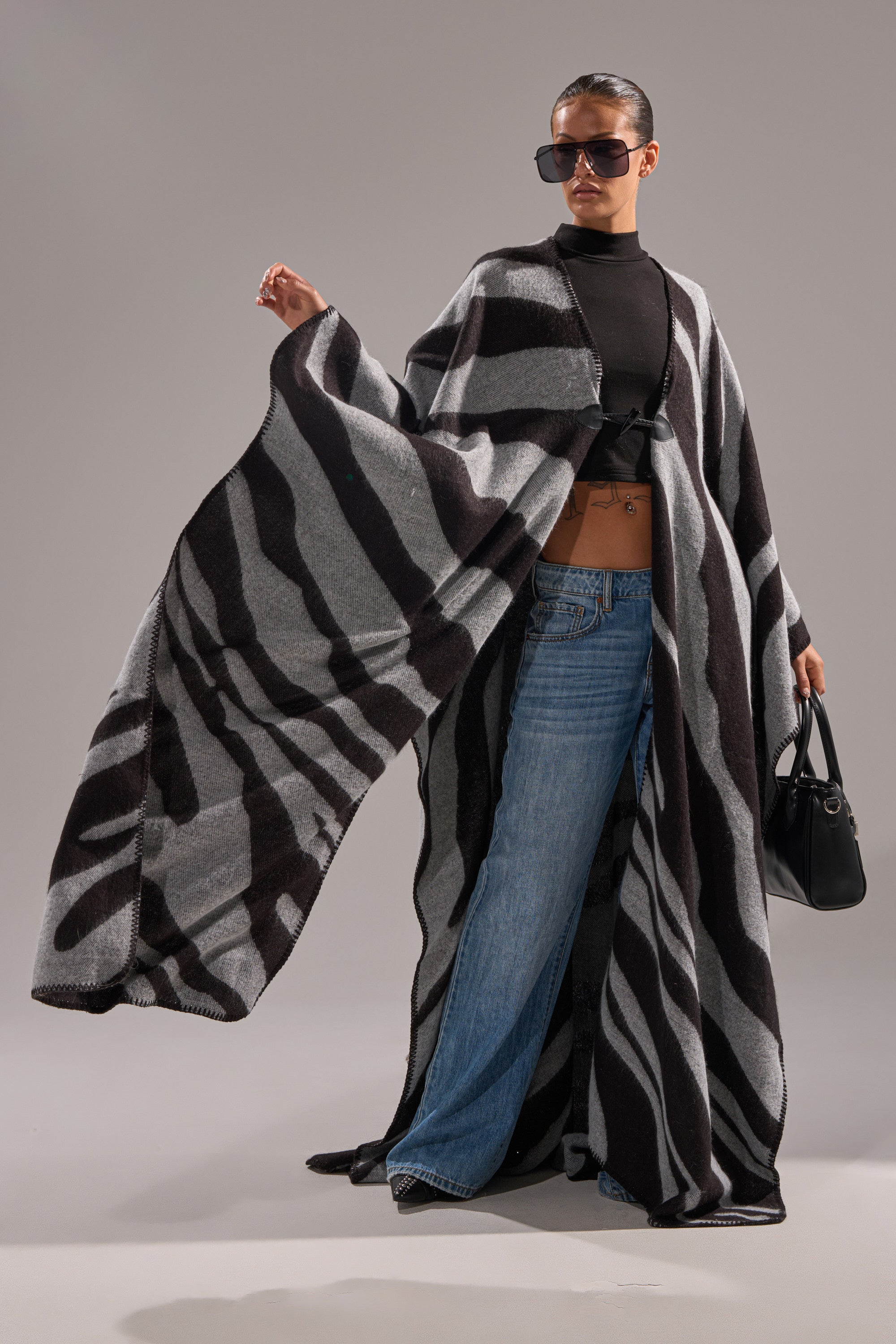Confidently posing while holding a black handbag, a person wears sunglasses, a black crop top, blue jeans, and the WILD ONE KNIT MAXI PONCHO—a long, flowing zebra-striped coat.