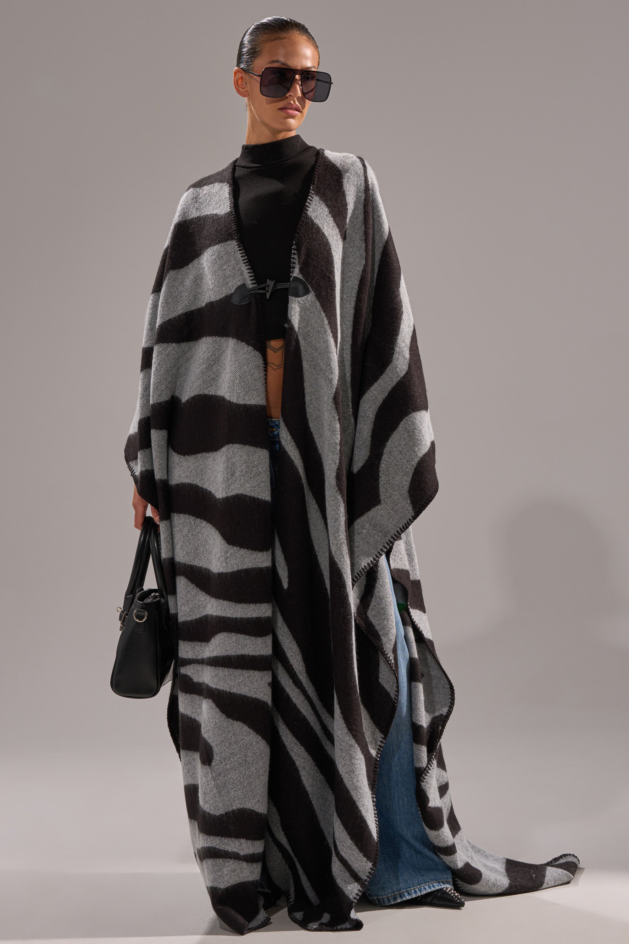 Wearing oversized sunglasses, a black top, blue jeans, and holding a black handbag, a person stands on a neutral background in the WILD ONE KNIT MAXI PONCHO, featuring a long, flowing zebra pattern.