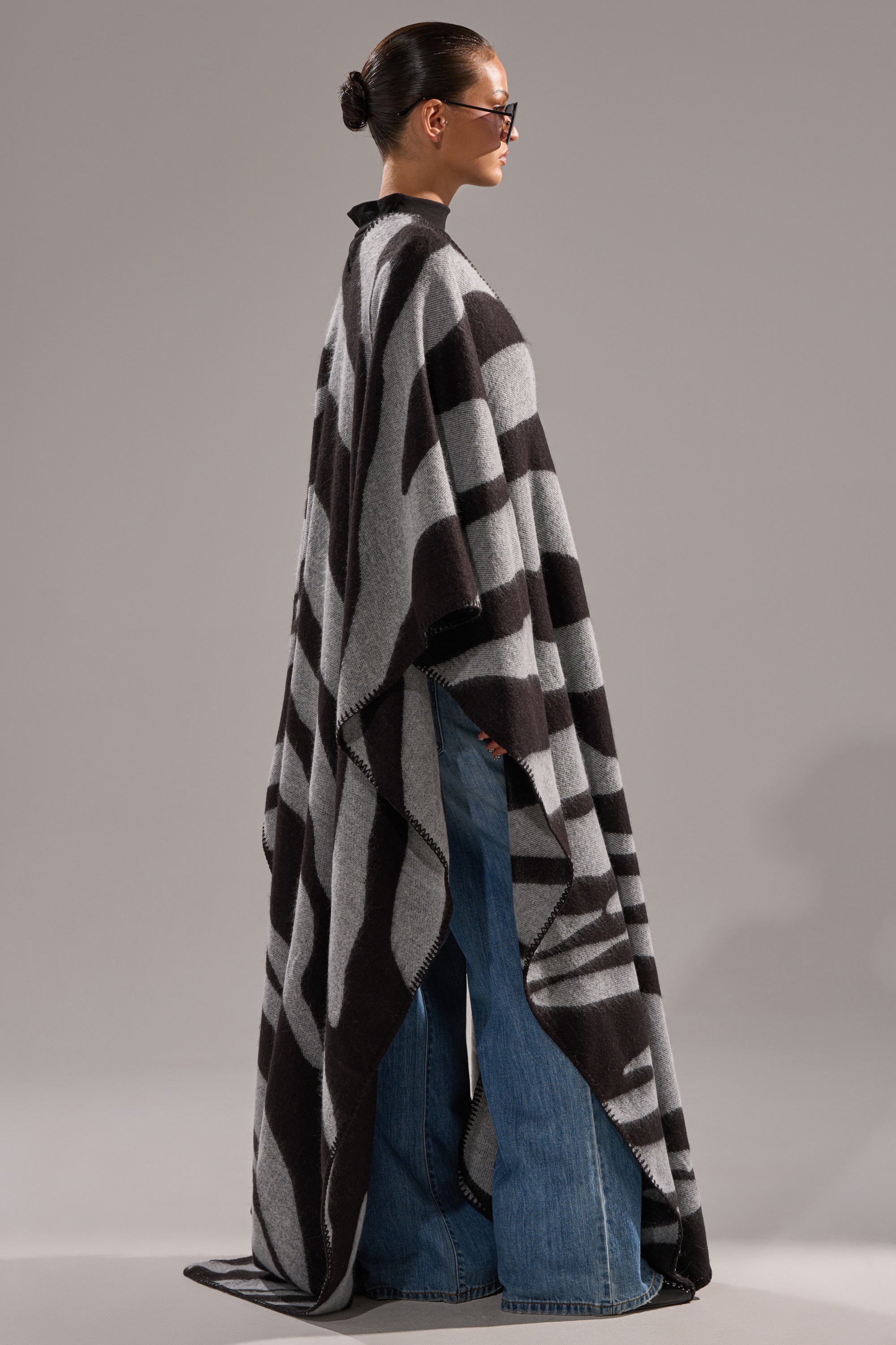 A person in sunglasses, blue flared jeans, and the WILD ONE KNIT MAXI PONCHO with a long black and gray geometric pattern, stands in profile against a plain gray background.