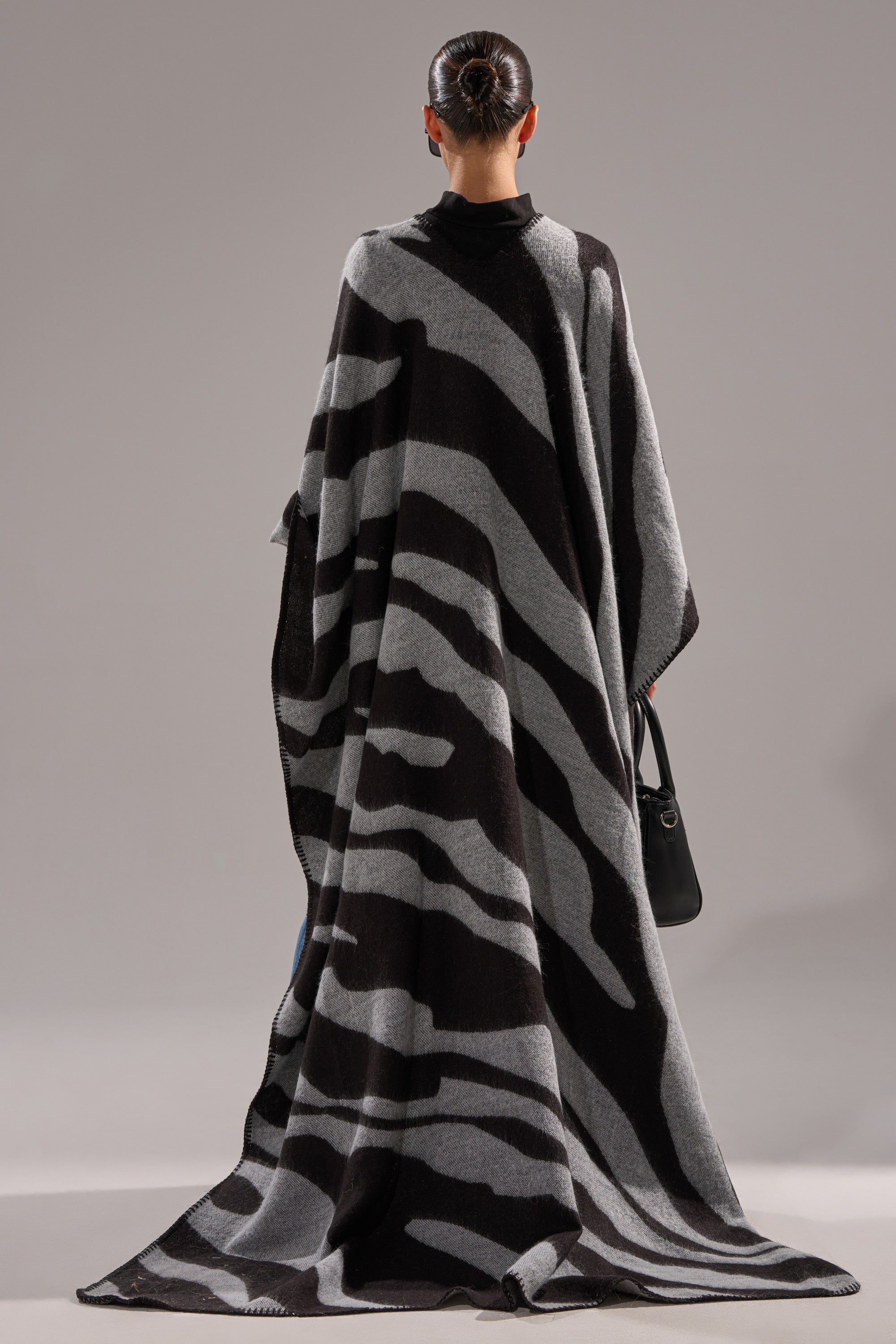 A person stands with their back to the camera, wearing the WILD ONE KNIT MAXI PONCHO in a black and gray zebra stripe that drapes to the floor. Their hair is in a neat bun, and they hold a small black handbag against a plain gray background.