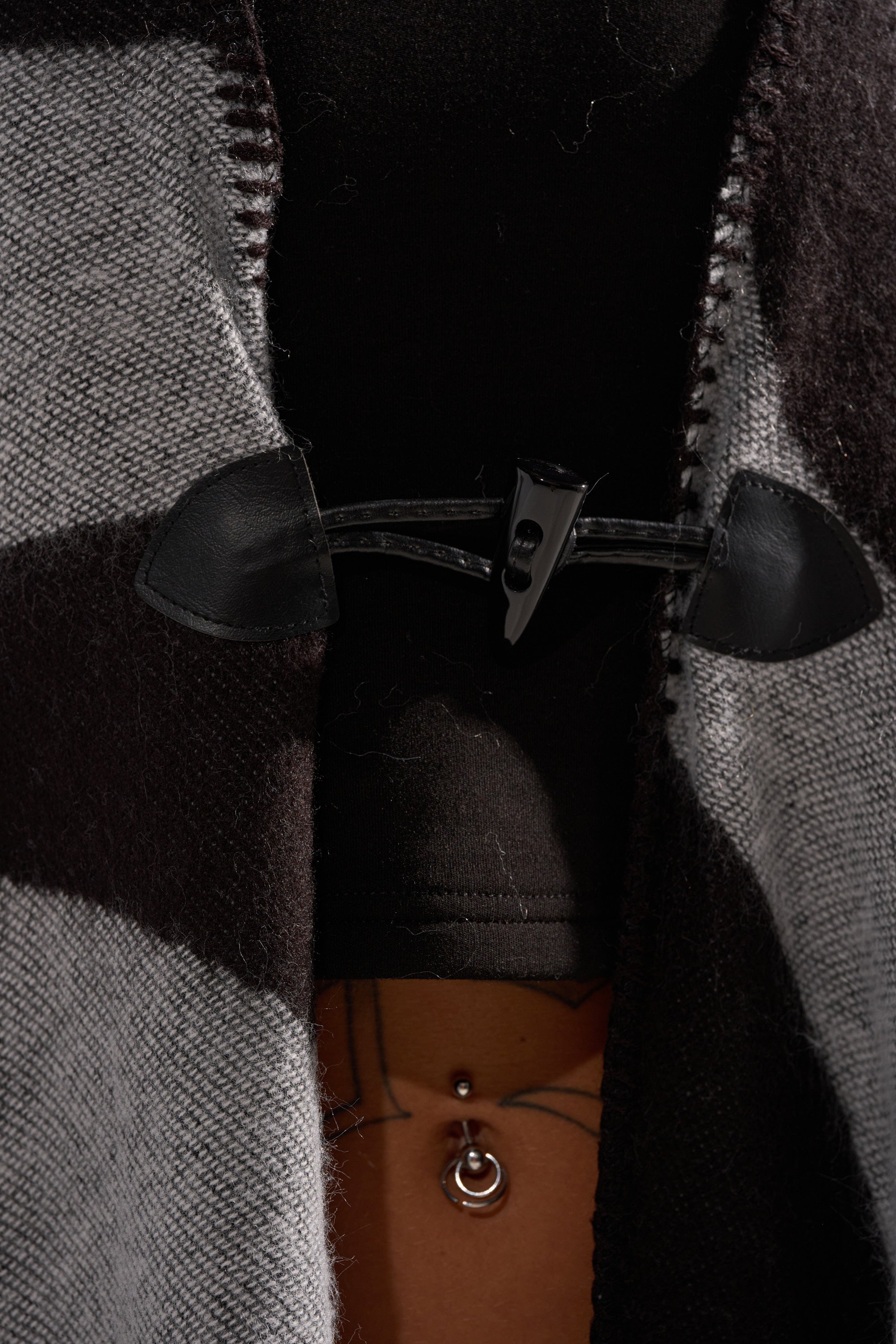Close-up of the WILD ONE KNIT MAXI PONCHO in black and grey, featuring a black toggle clasp. A glimpse of a bare midriff with a belly button piercing and part of a tattoo is visible beneath the poncho.