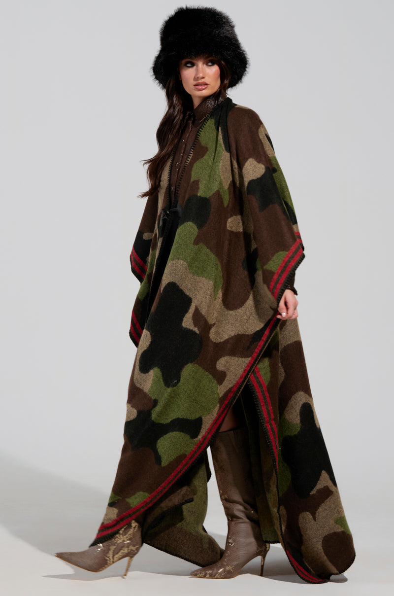 A person confidently stands in a flowing CAMO NIGHTS MAXI PONCHO crafted from ultra-soft knit, paired with a black fur hat. Knee-high boots with a slight heel complete the ensemble, while earthy tones of green, brown, and beige accented by red breathe life into the camo print pattern.