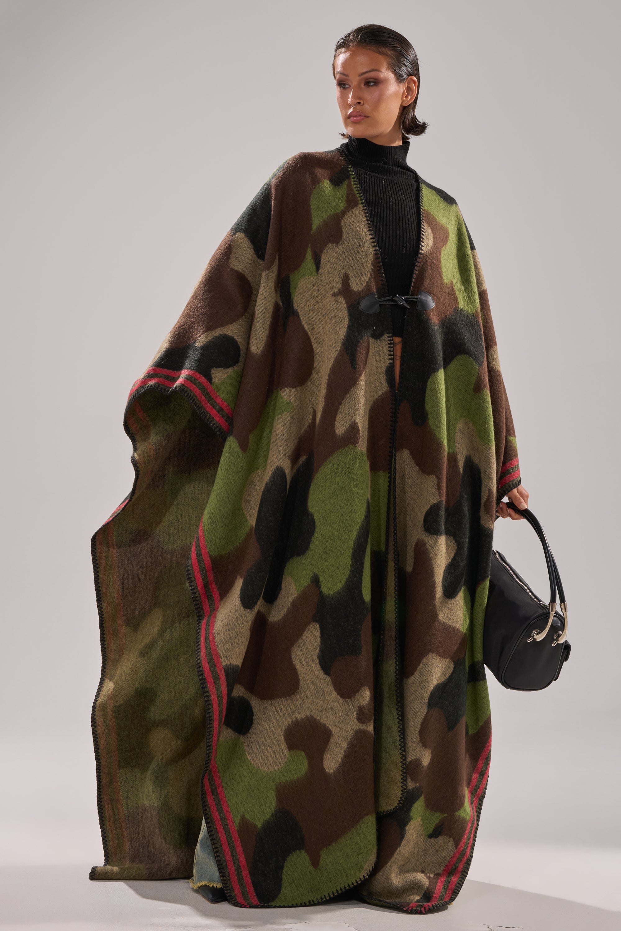 A woman wears the CAMO NIGHTS MAXI PONCHO, featuring a flowy camo design with red striped edges over a black outfit. She holds a black handbag and gazes confidently to the side against a plain background.