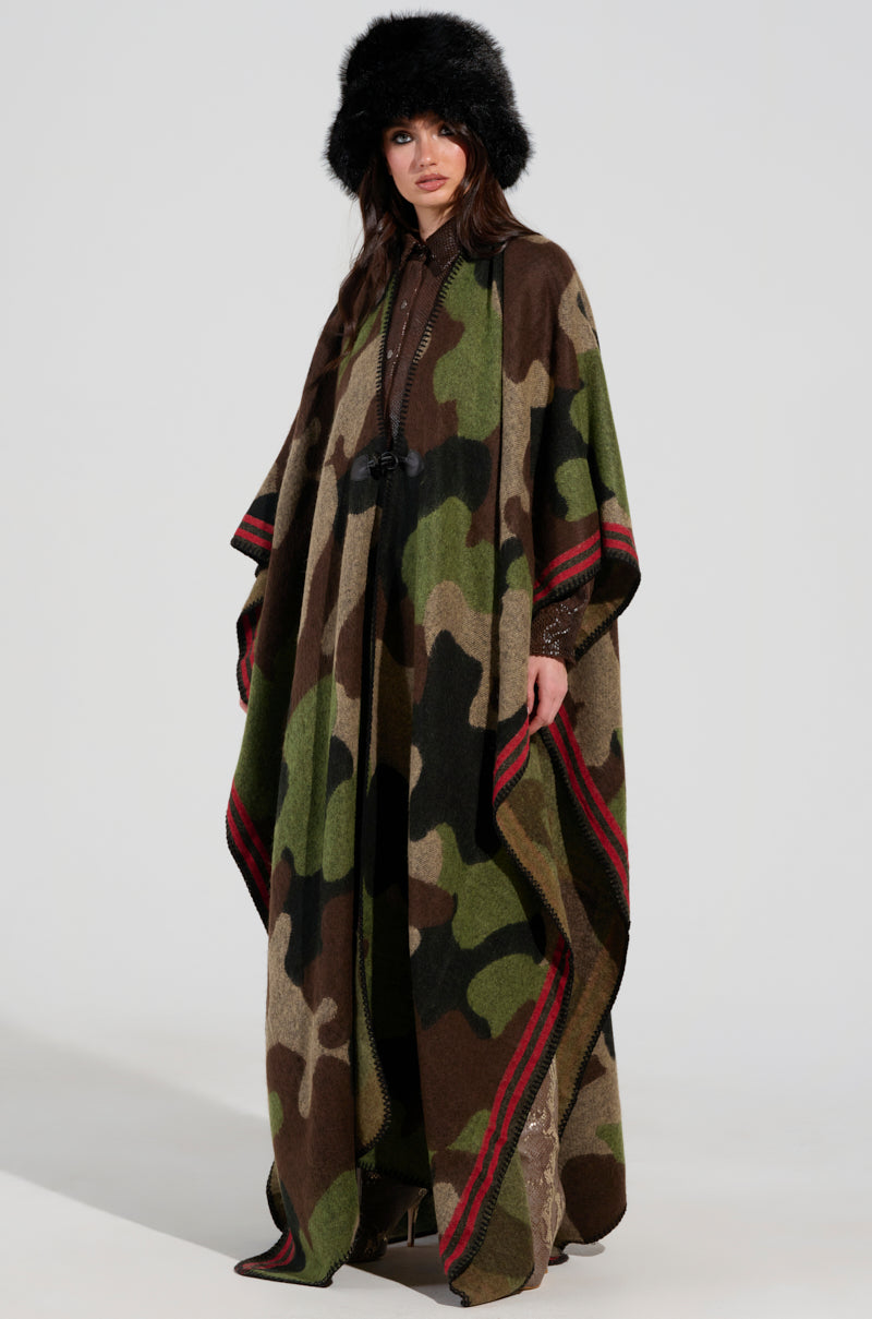 Wearing the ultra-soft knit CAMO NIGHTS MAXI PONCHO, a person stands against a plain background. Their long hair and black fur hat accentuate the stylish, outdoorsy look that seamlessly blends camo print with red-and-black stripes.
