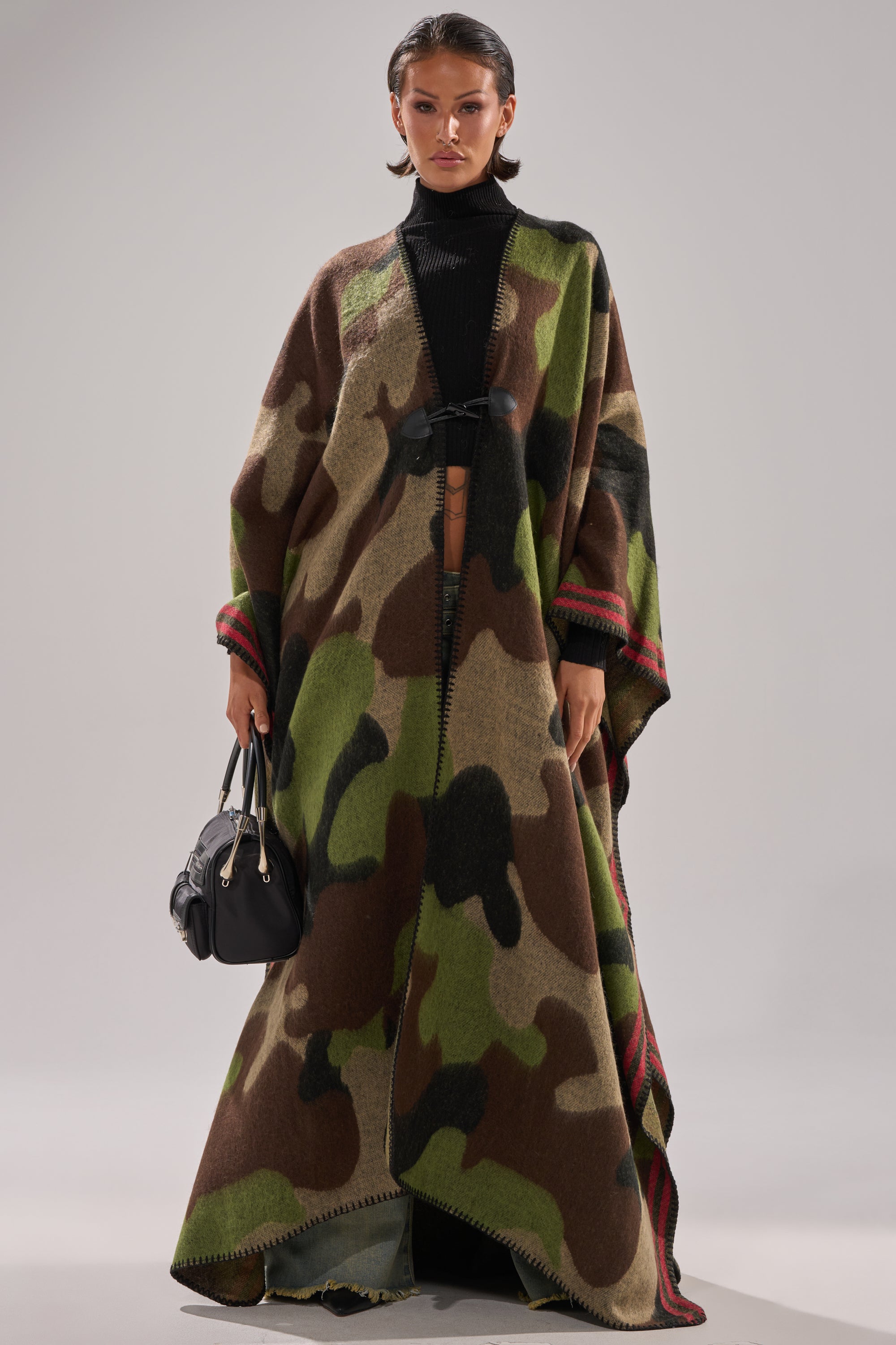 Wearing the CAMO NIGHTS MAXI PONCHO with red accents over a black turtleneck and holding a black handbag, a person stands against a plain light gray background.