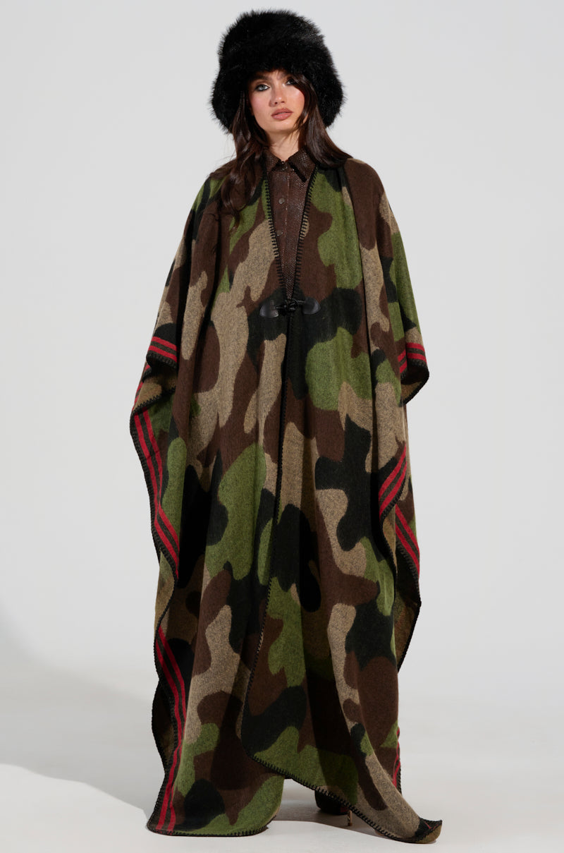 A person is dressed in the CAMO NIGHTS MAXI PONCHO, which showcases a camouflage print with red stripe accents. They stand facing forward, wearing a dark fur hat and having long hair. With a serious expression, they pose against a plain gray backdrop.