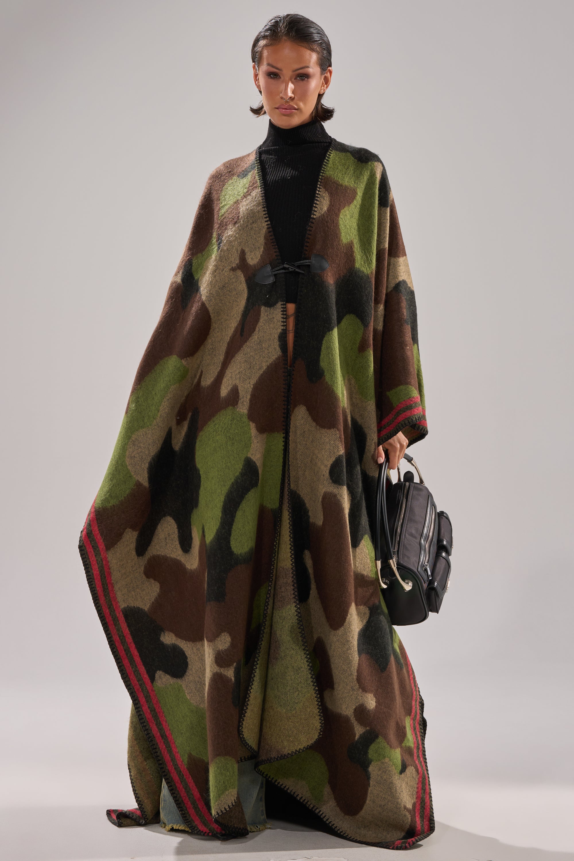 Wearing the CAMO NIGHTS MAXI PONCHO with red trim over a black turtleneck, a woman holds a black handbag and gazes confidently at the camera against a simple background.