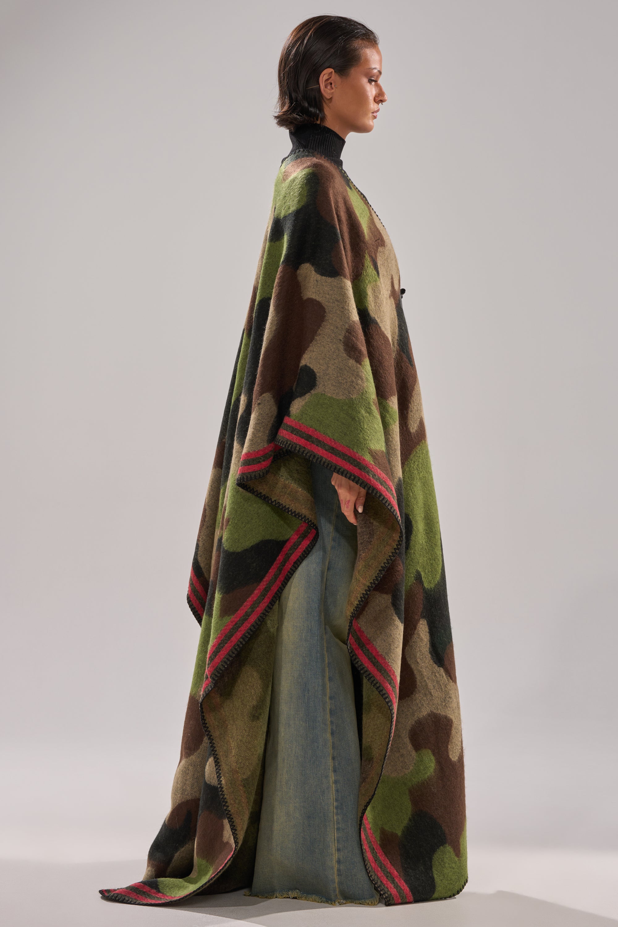 A person stands in profile wearing the CAMO NIGHTS MAXI PONCHO, a flowy camo piece with red-striped edges, layered over wide-legged light blue pants. The background is plain and neutral.