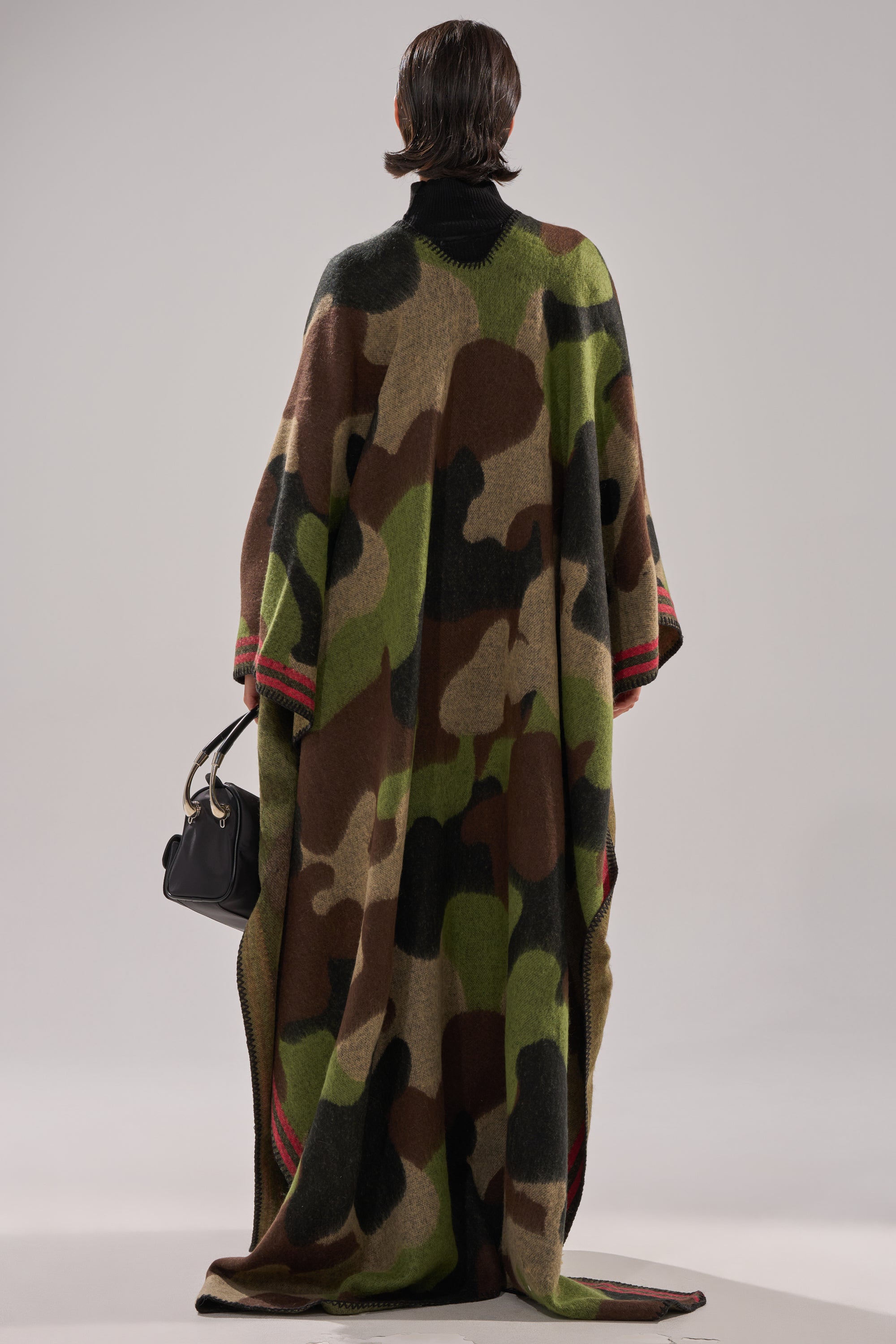 A person with short dark hair, seen from behind, wears the CAMO NIGHTS MAXI PONCHO with red accents and holds a black handbag. The background is plain and neutral.