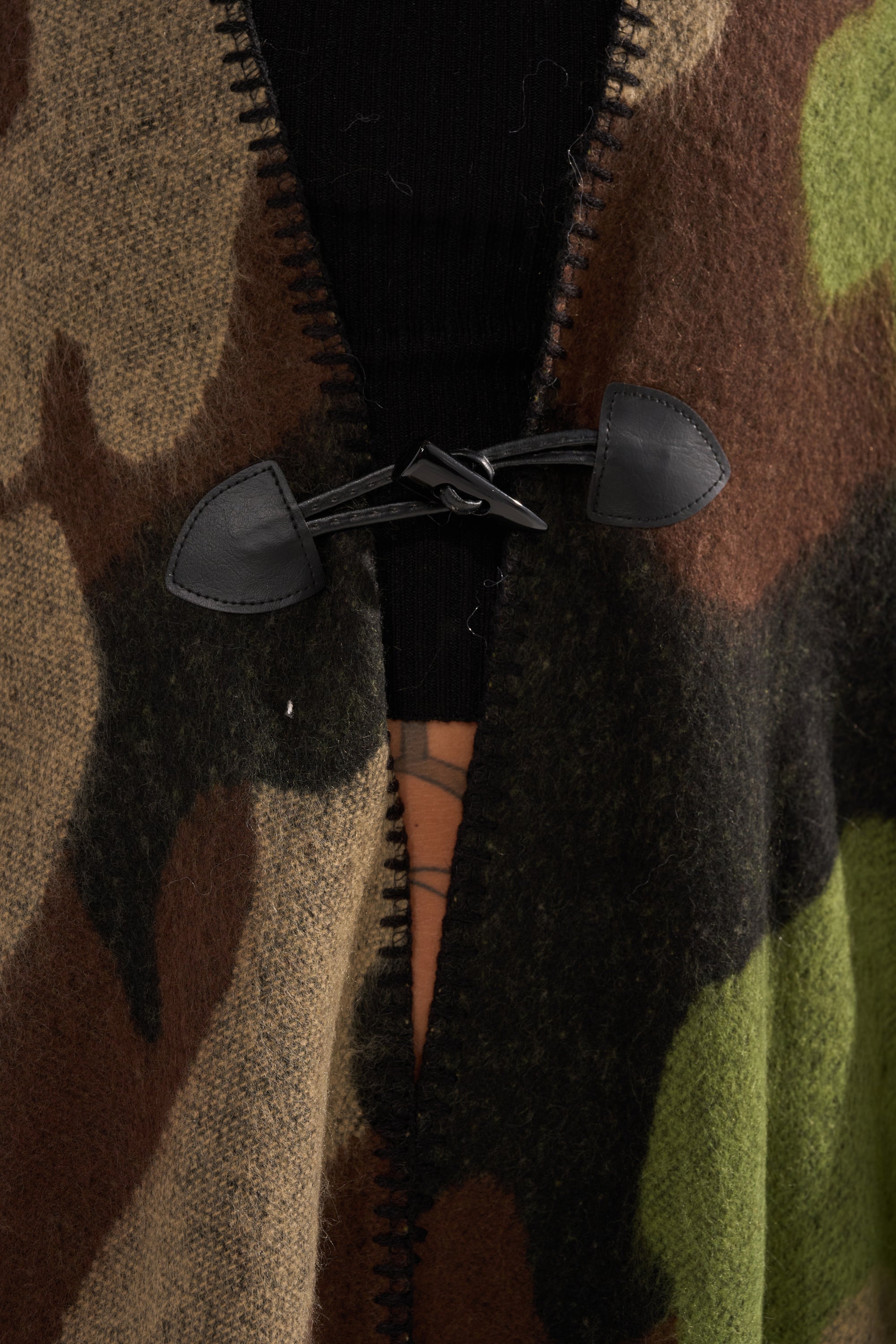 Close-up of the CAMO NIGHTS MAXI PONCHO, featuring a flowy camo design with a black toggle clasp at the center. The zipper opening reveals a black garment underneath.
