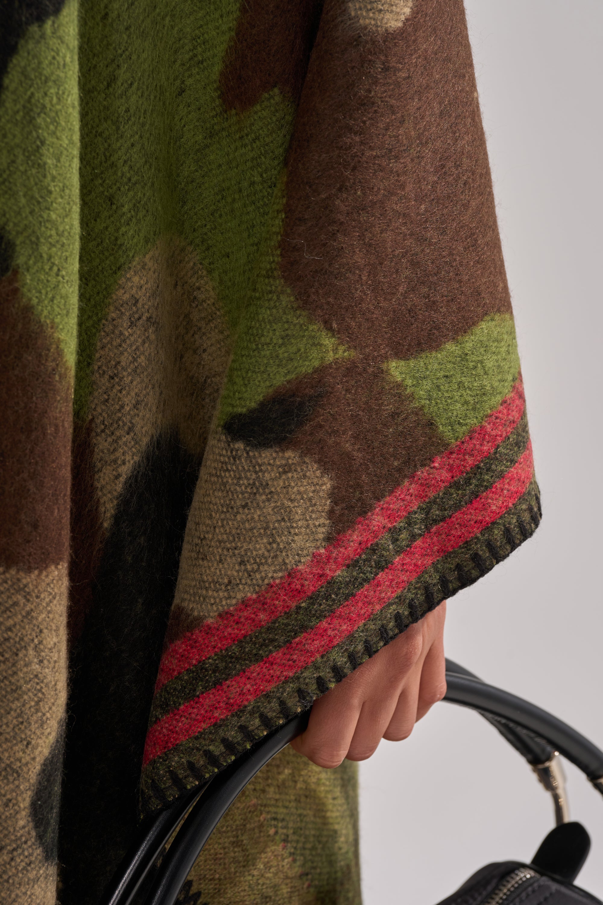 A close-up of a hand holding a black handle, wearing the ultra-soft CAMO NIGHTS MAXI PONCHO featuring green and brown camouflage with red stripes along the edge.