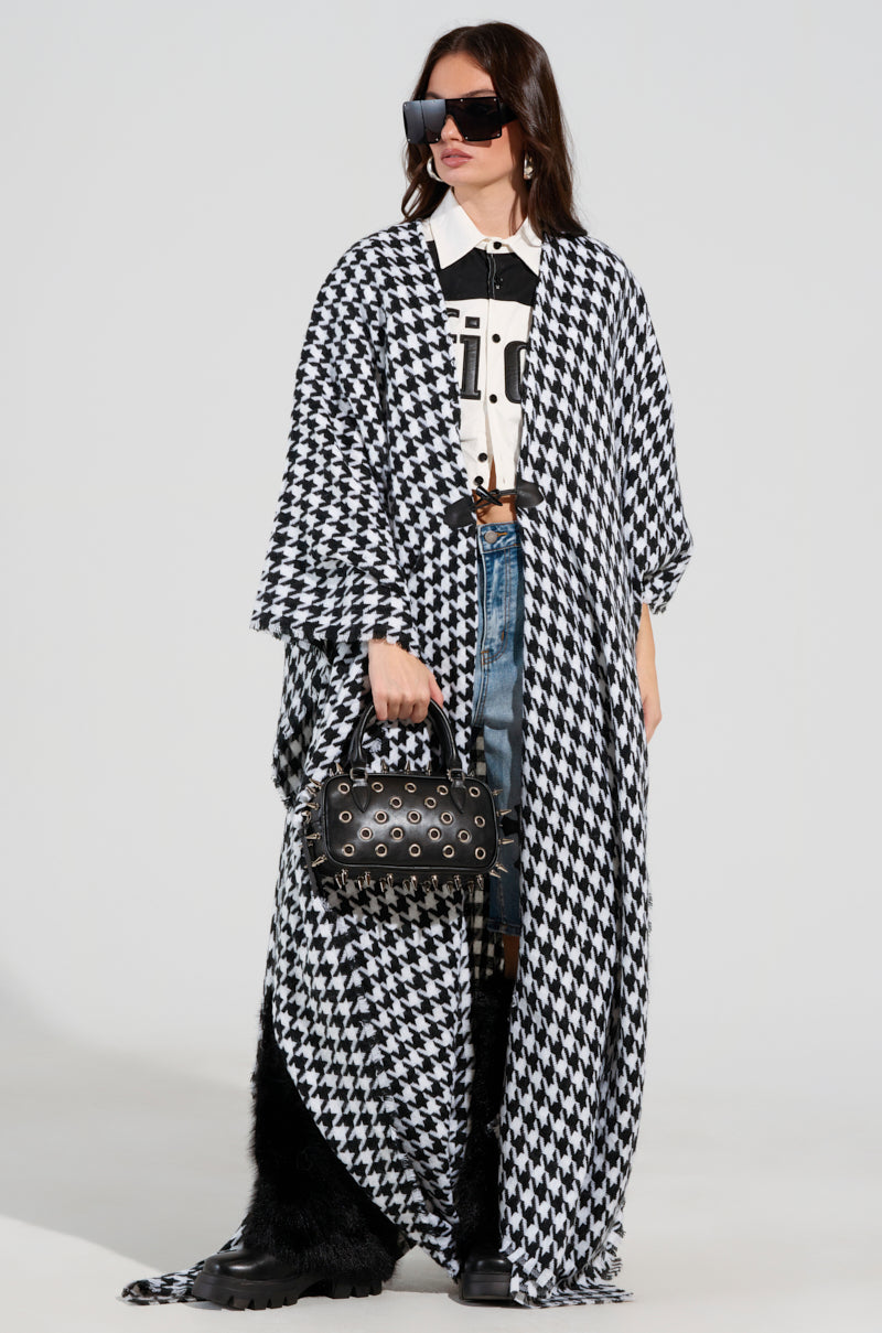 A person is dressed in the ELEVATED LIFE HOUNDSTOOTH PONCHO, paired with black sunglasses and carrying a black studded bag. They showcase a white shirt with black lettering underneath, complemented by blue jeans and striking black furry boots. 0