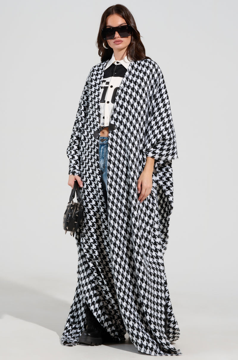 A person wearing large black sunglasses and an oversized silhouette showcases the ELEVATED LIFE HOUNDSTOOTH PONCHO, crafted from a cozy knit fabric, over a black and white top and blue jeans. They hold a small black handbag while standing confidently against a plain background. 1