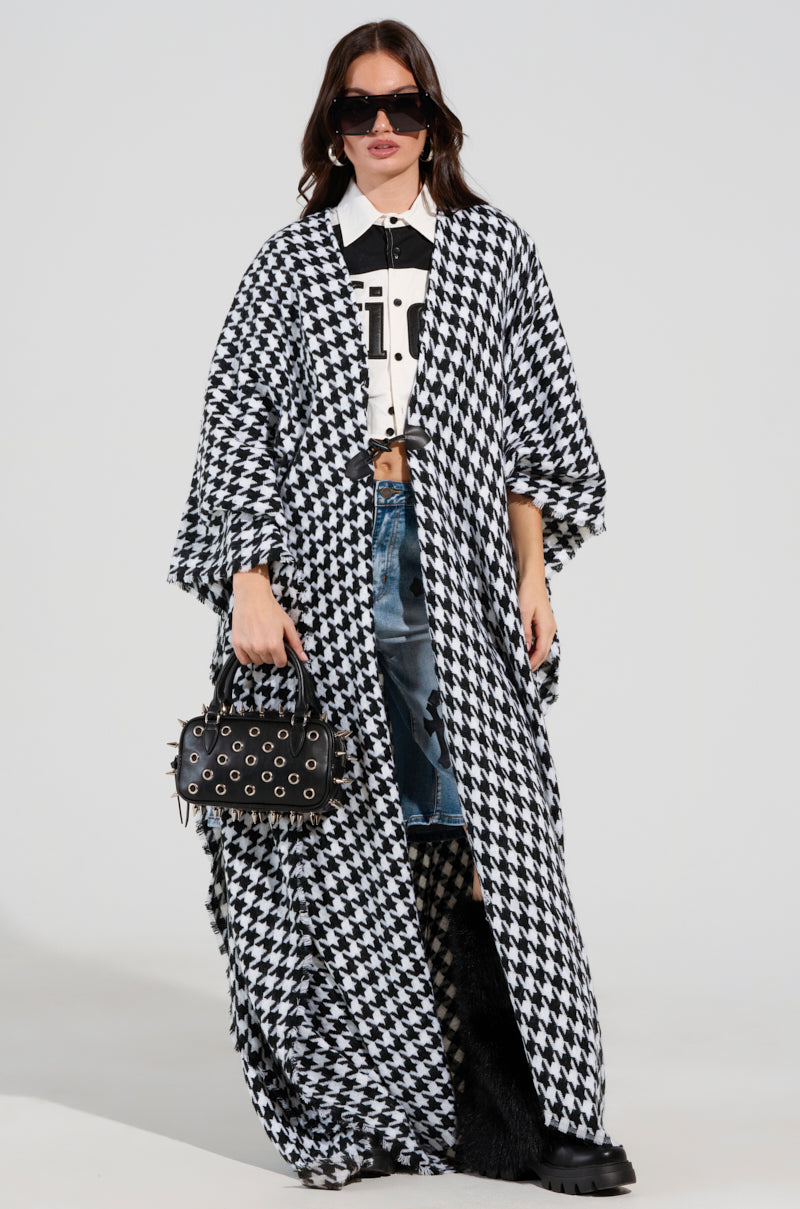 Wearing the ELEVATED LIFE HOUNDSTOOTH PONCHO, a person exudes style with an oversized silhouette over a top and denim skirt. Their long dark hair cascades under chic sunglasses, and they carry a studded black handbag while sporting sleek black shoes. 2