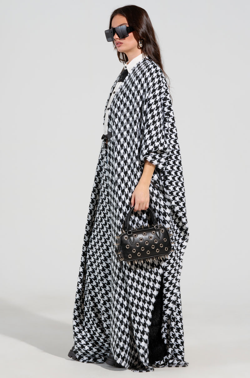 A woman in an oversized silhouette wears the ELEVATED LIFE HOUNDSTOOTH PONCHO over a white collared shirt, accessorized with oversized sunglasses and a small black handbag adorned with studs. 3