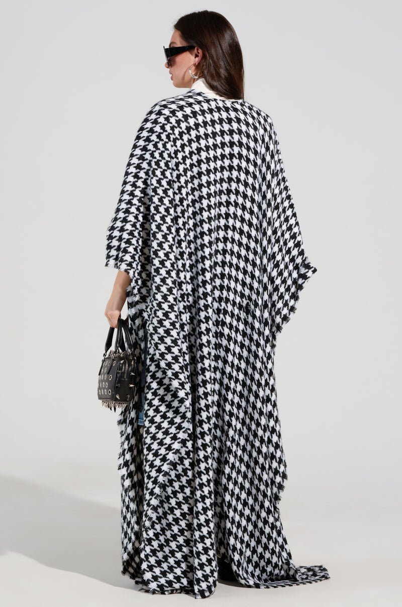 A person with long dark hair stands with their back to the camera, showcasing the ELEVATED LIFE HOUNDSTOOTH PONCHO. They hold a studded handbag and wear large sunglasses, completing the chic look against a plain, light-colored background. 4