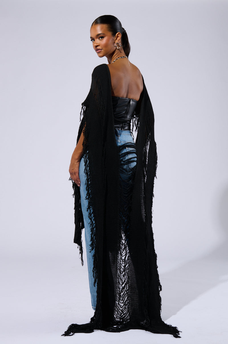 A woman stands confidently against a plain background, wearing a black strapless top, distressed light blue jeans, and the GOOD VIBES DISTRESSED PONCHO IN BLACK draped over her shoulders. She has slicked-back hair and gold hoop earrings, epitomizing a Boho Chic ensemble as she looks back over her shoulder.