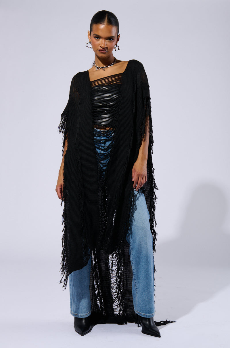 A person poses against a plain backdrop, showcasing the GOOD VIBES DISTRESSED PONCHO IN BLACK, effortlessly layered over a black leather top and blue jeans. They accessorize with a spiked choker and star-shaped earrings, exuding boho chic vibes. With a confident expression and slicked-back hair, they command attention effortlessly.