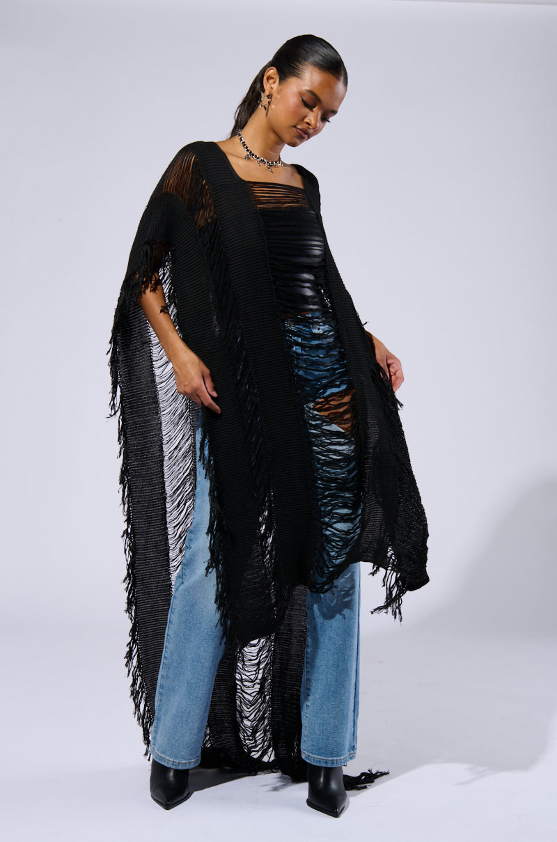 A person wearing the GOOD VIBES DISTRESSED PONCHO IN BLACK over a fitted black top and light blue jeans. Sporting dark hair pulled back, they strike a relaxed pose against a gray background. With black boots and a necklace completing their Boho Chic Ensemble, they exude effortless cool.
